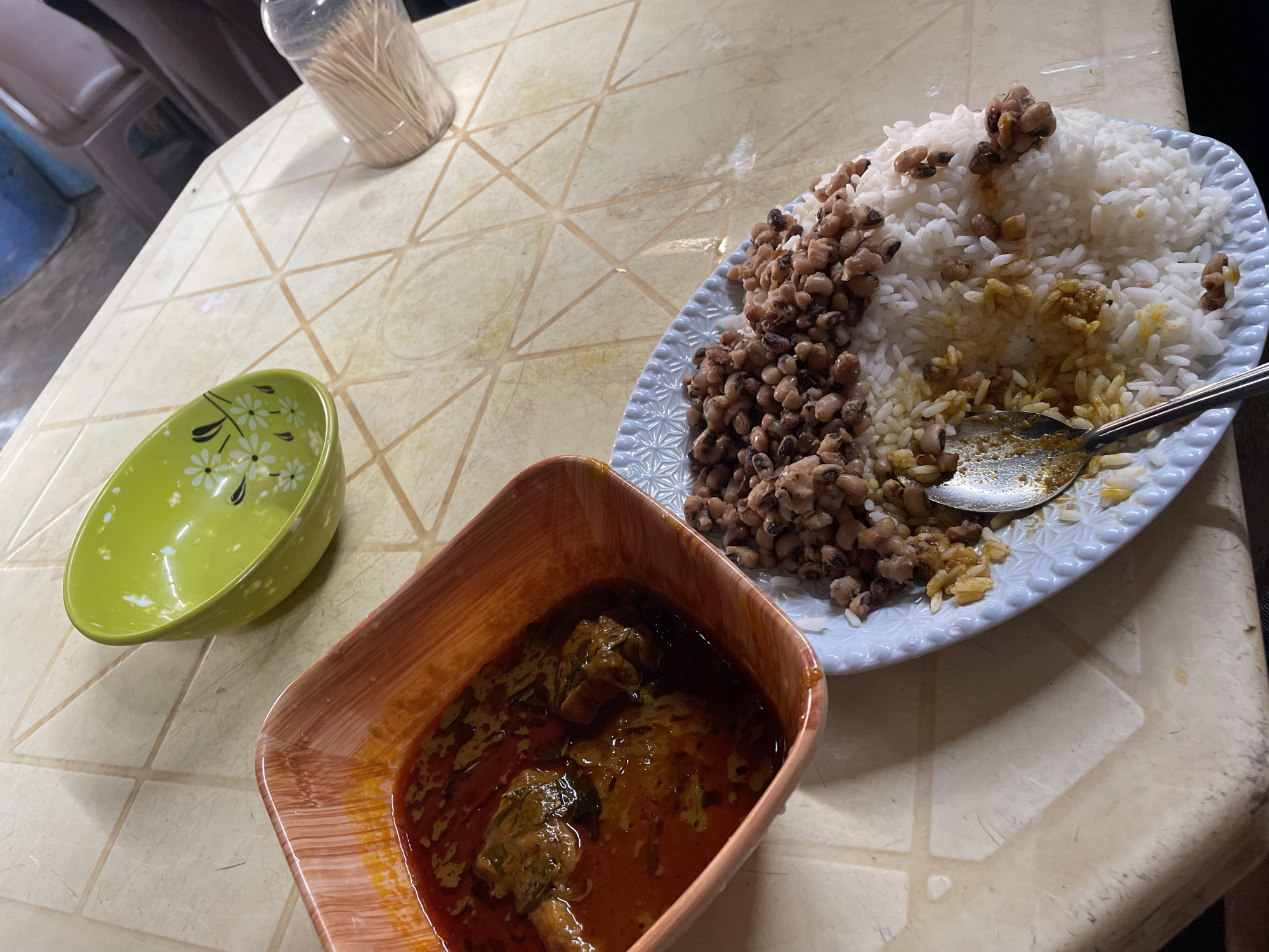 On a patterned, off-white table, likely indoors, a plate of white rice and brown beans is positioned next to a square bowl filled with a red-brown curry. A green bowl with white floral patterns sits to the left of the curry. A stainless steel spoon rests on the rice and beans. A plastic container filled with toothpicks is present in the background. The image suggests a meal is being served, possibly in a residential or commercial setting. The location is Obosi, Nigeria. No people are visible in the image.