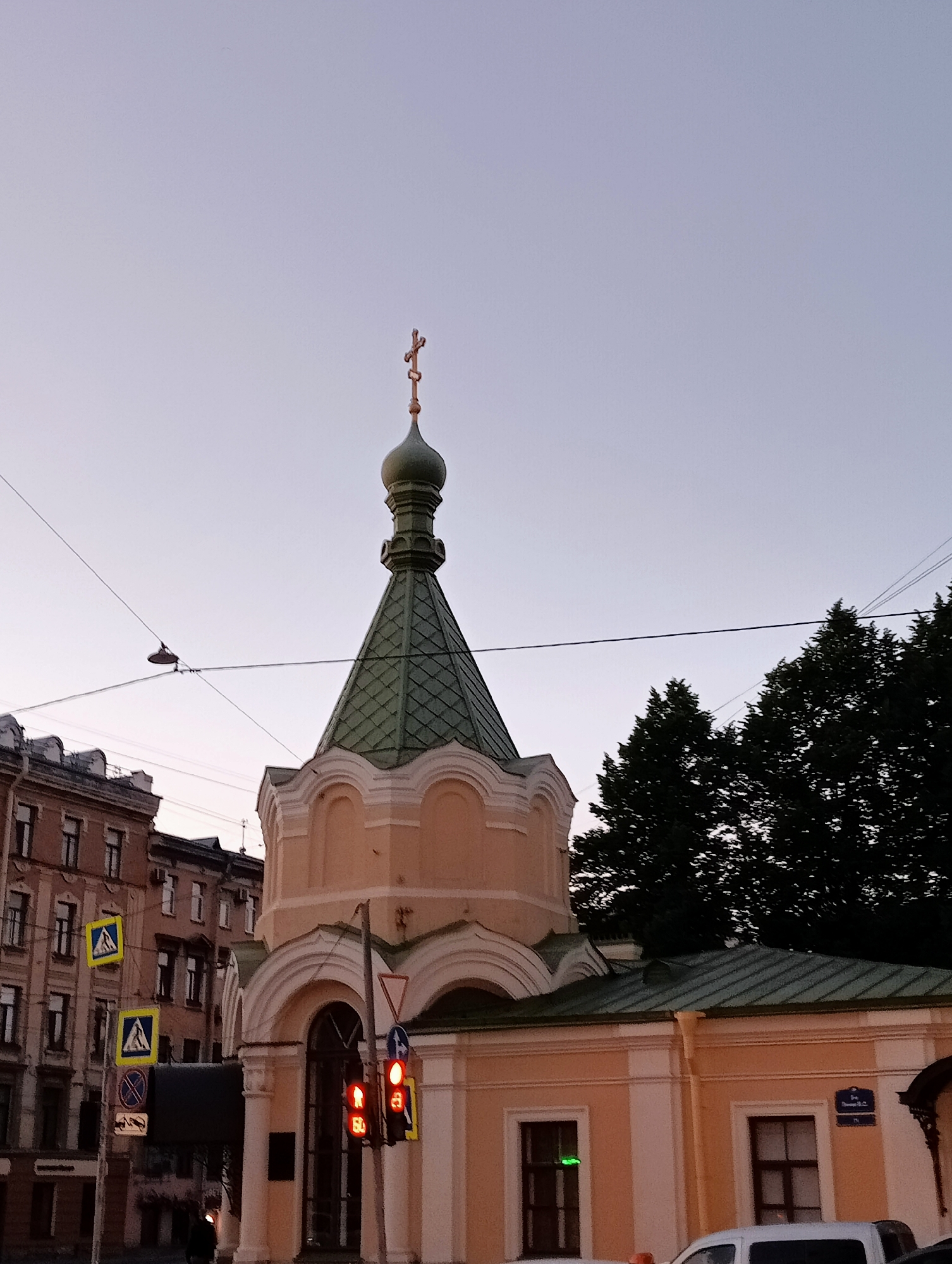 The image captures a street scene in Saint Petersburg, Russia, at dusk. The prominent subject is the green-domed chapel with a cross at its apex, positioned in the center right of the frame. It is a light-peach colored building with white decorative molding. To the left, a multi-story building with a beige facade and numerous windows is visible, characteristic of St. Petersburg architecture. Several traffic signs are present, including yellow diamond-shaped signs with pedestrian symbols and a red circular sign prohibiting stopping. A traffic light with red and yellow illuminated bulbs is also visible, indicating traffic control. Overhead, electrical wires crisscross the sky, and a street lamp hangs suspended between them. Silhouetted trees are present on the right side of the image, and a dark-colored car is parked in the foreground on the right. The sky is a pale, twilight blue.