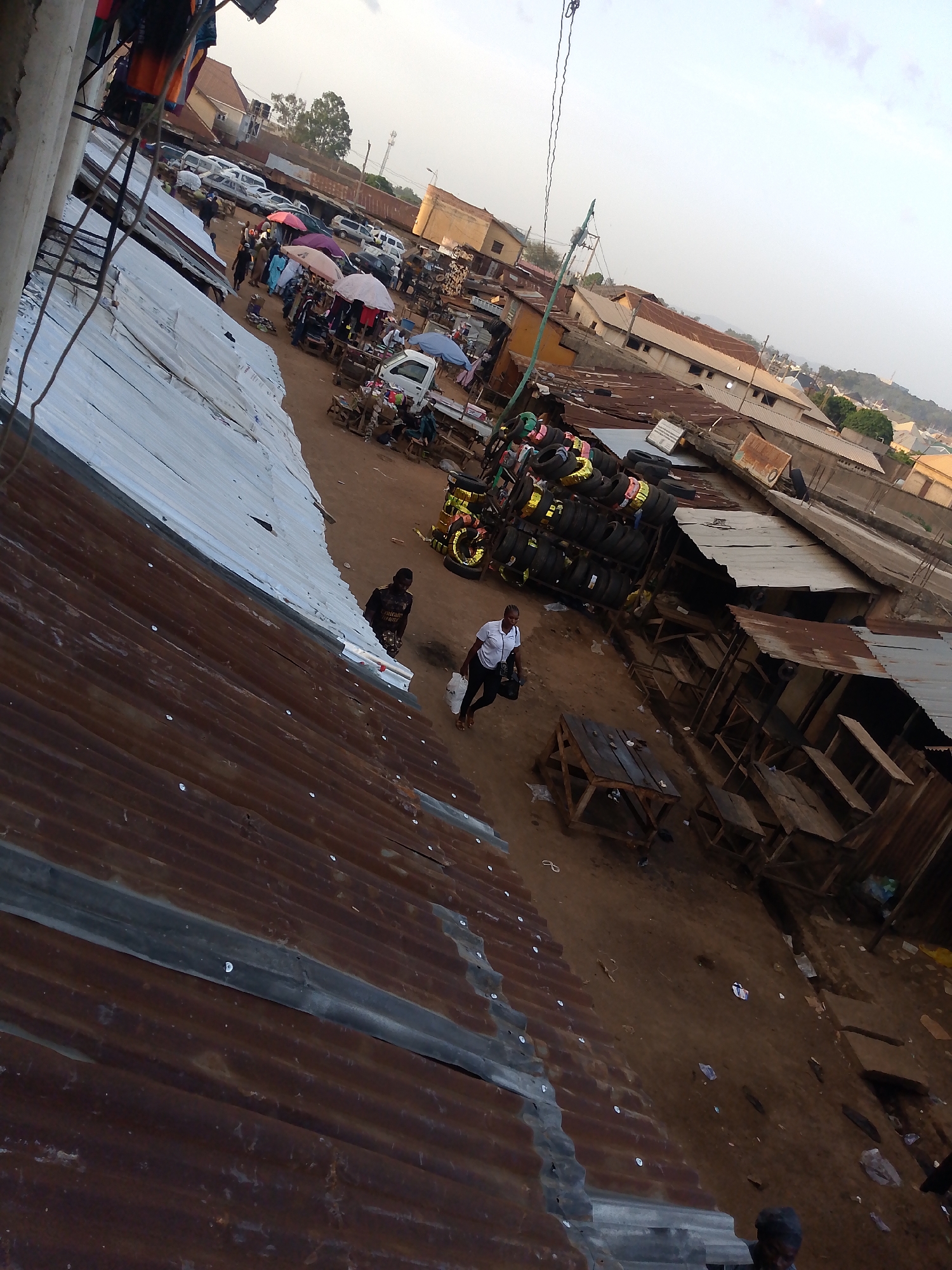 The image depicts an outdoor market scene in Bukuru, Nigeria, viewed from an elevated angle. The foreground is dominated by rusted corrugated metal roofs. A dirt path runs through the center of the frame, bordered by wooden stalls and shops. Several people are visible; one woman in a white shirt is walking along the path, carrying a bag. A stack of tires sits near the center of the frame. In the background, there are more stalls and shops, with cars and people clustered together. Umbrellas provide shade for vendors and customers. The sky is overcast.