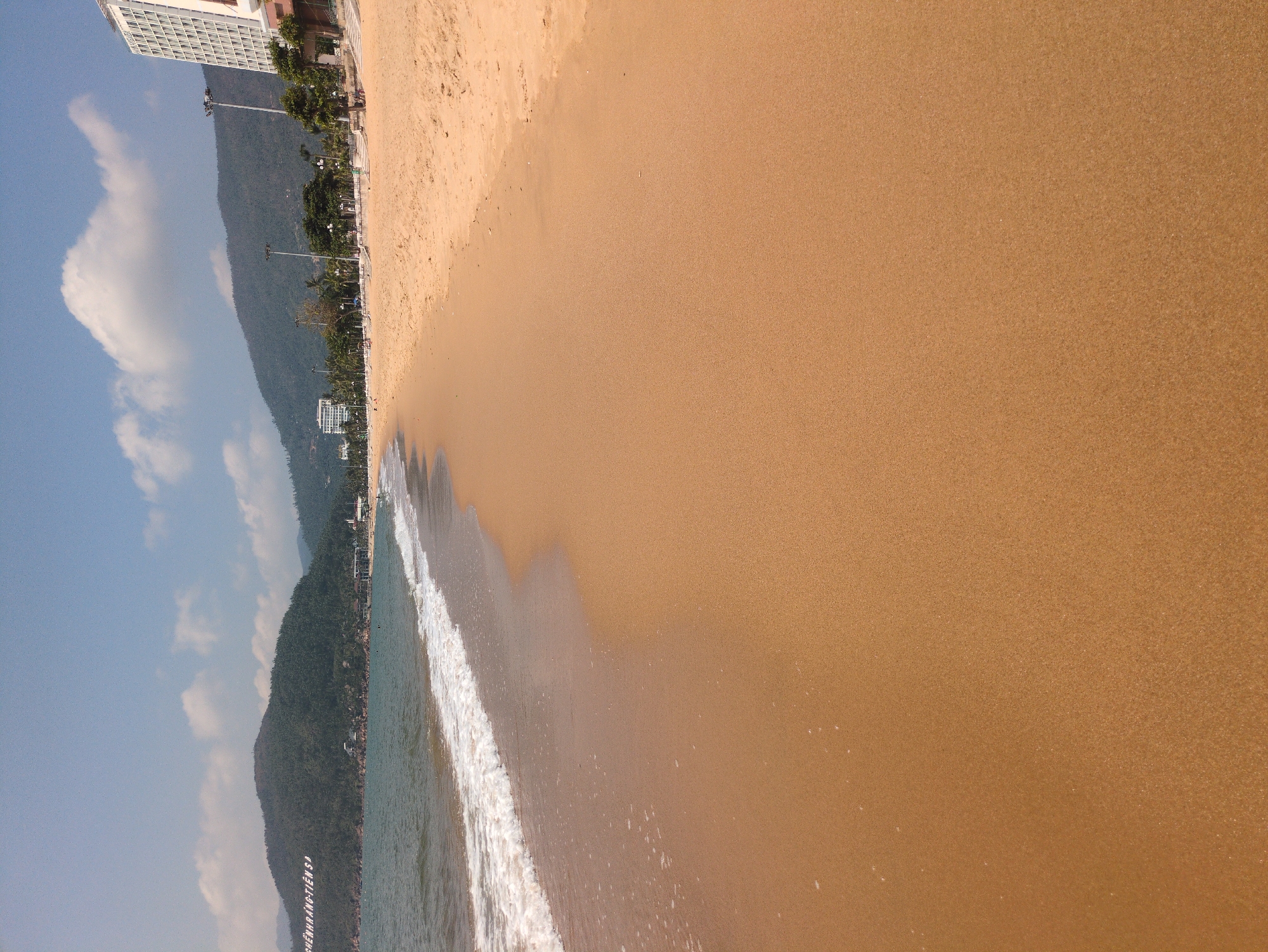 The image depicts a coastal scene in South Quy Nhon Ward, Vietnam. A sandy beach occupies the majority of the frame, with the ocean visible along the left edge. Gentle waves are breaking on the shore. A line of vegetation and buildings, including a tall white structure, runs parallel to the beach. Mountains rise in the background, partly obscured by clouds in the sky.