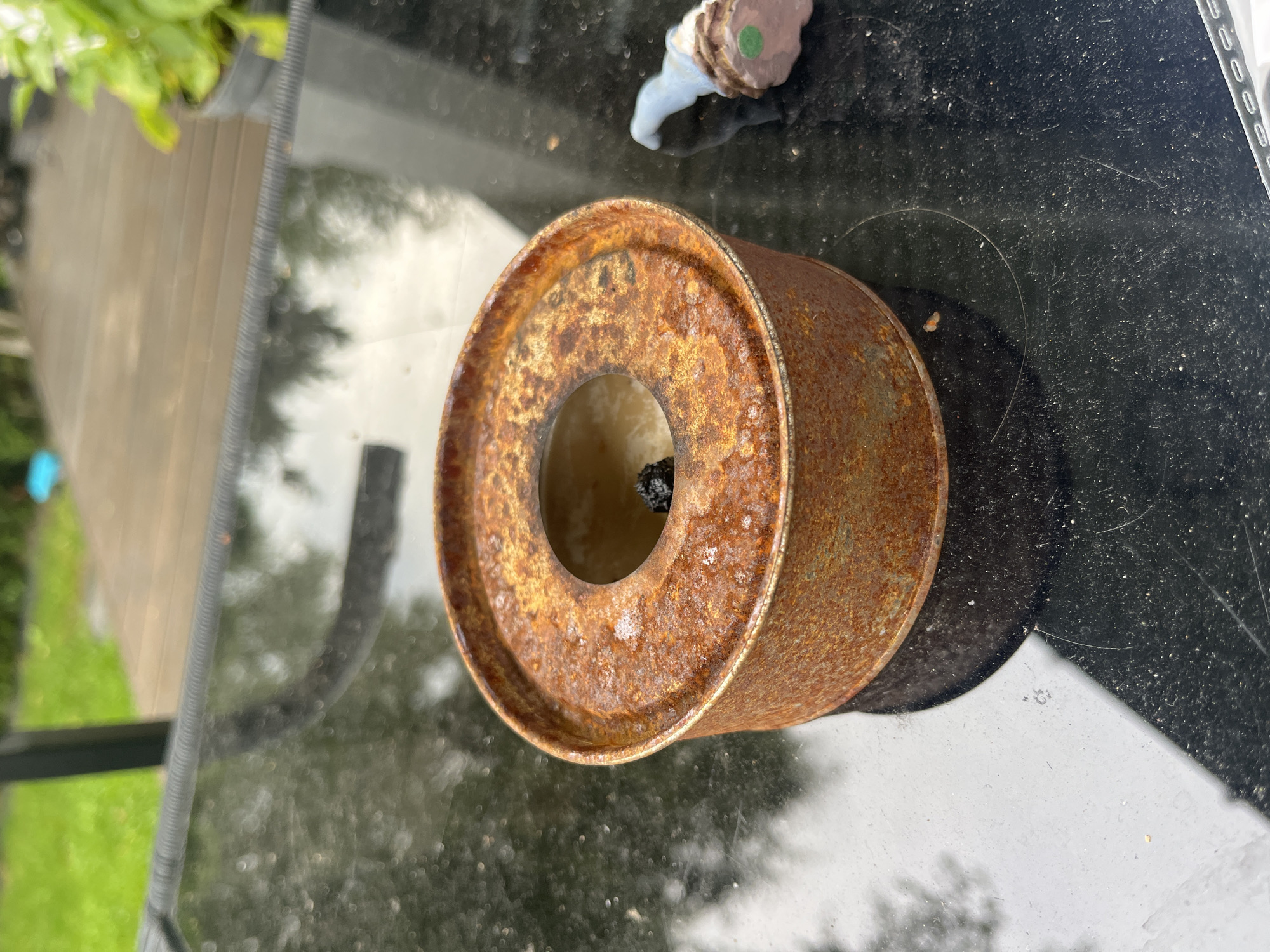 A rusty, circular metal tin with a hole in the center sits on a black glass table. Inside the hole, a small black object is visible. To the right of the tin is a small, decorative gnome with a blue hat and brown base. The table is outdoors, with greenery and a wooden deck visible in the background. The overall impression is of a backyard setting in Haabneeme, Estonia.