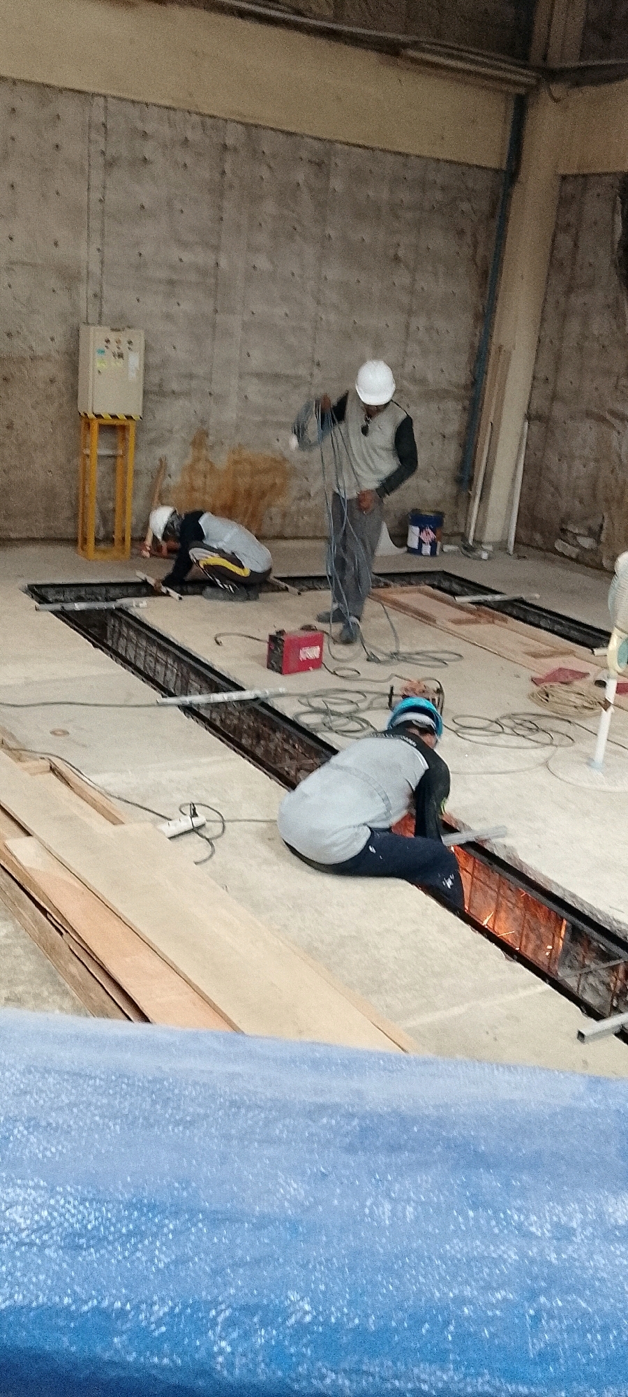 In Depok, Indonesia, construction is underway within a concrete building, indicated by the raw concrete walls and floors. Three individuals, dressed in construction attire including hard hats and safety vests, are actively engaged in work. One person, positioned in the foreground, is kneeling and welding within a trench, sparks emanating from the work. To the left, another worker is also kneeling, appearing to be involved in some form of installation or repair within a similar trench. A third individual stands further back, holding a bundle of electrical cords, possibly involved in setting up or managing the electrical supply for the work. Various construction materials, including lumber and electrical equipment such as a red welding machine and a white power strip, are visible around the site. The environment suggests an ongoing construction project, possibly involving structural reinforcement or infrastructure installation within the building.