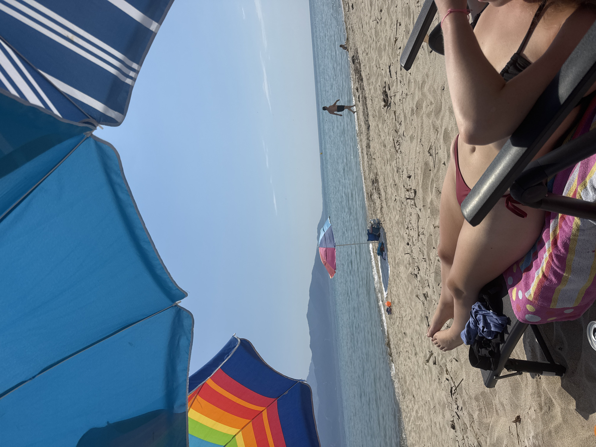 This image captures a first-person perspective of a beach scene in Unknown, Unknown, under a clear blue sky. In the immediate foreground, a person is relaxing on a black beach chair, viewed from the waist down. Their legs are crossed at the ankles, and they are wearing a dark red bikini bottom and a black bikini top. A pink, yellow, and white striped towel covers the chair beneath them. A crumpled blue garment and a black piece of clothing rest on the chair near their feet. A silver can is partially visible in the bottom left corner.
Above, two large beach umbrellas dominate the upper portion of the frame: a plain blue one is central to the left, and a blue and white striped one is to the far right. A colorful rainbow-striped umbrella is partially visible below the plain blue one on the left.
The midground features a sandy beach meeting a calm body of water, likely the sea. Scattered driftwood and debris are visible on the sand. In the shallow water near the shore, an adult male in dark swim trunks walks to the right. Further down the beach, a small child is visible at the water's edge. A small, multi-colored (pink, light blue, white) beach umbrella is pitched on the sand to the left of the walking man, with some blue items and a cooler or bag behind it. An orange bucket or toy and some footwear are on the sand near the water. A yellow buoy floats in the water in the mid-distance.
Across the water, a range of hazy, dark blue mountains forms the horizon line. The sky above is cloudless and bright blue.