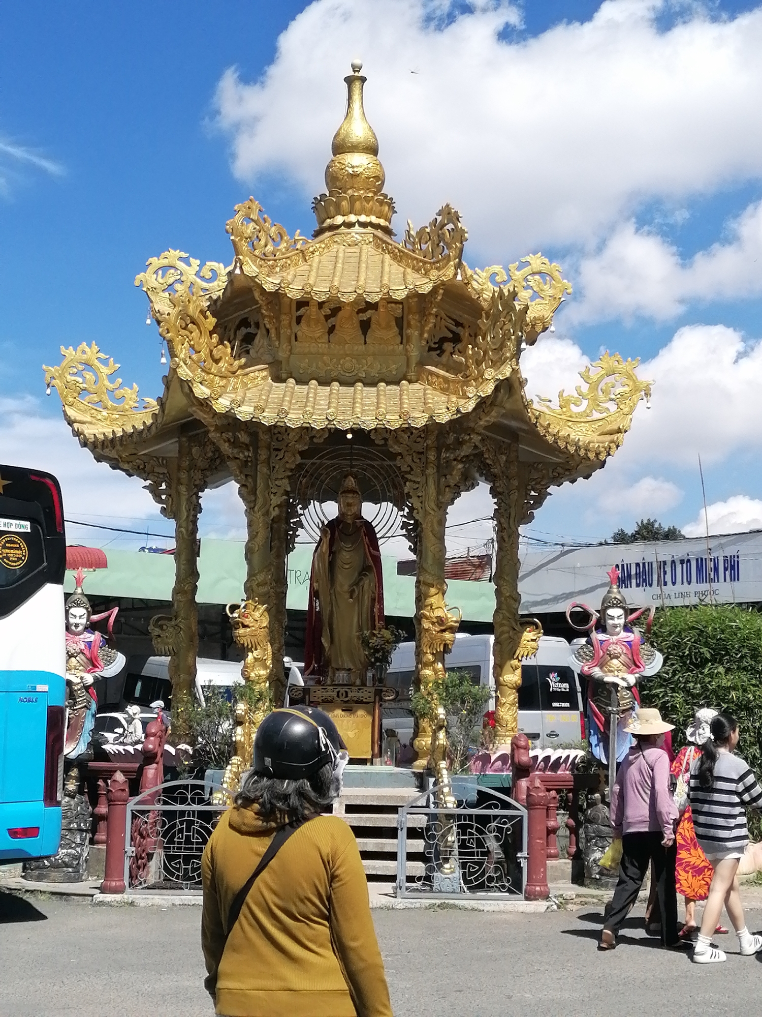 The image depicts a Buddhist temple in Xuân Thành, Vietnam. The main subject is a golden pagoda adorned with intricate carvings and statues. A large, standing golden statue of Buddha is prominently displayed within the pagoda. Flanking the pagoda are two warrior statues.
In the foreground, a person wearing a yellow sweater and a black helmet is seen from behind, looking towards the temple. To the right, three individuals are walking away from the pagoda. The woman in the center wears a pink shirt and a straw hat, while the woman on the far right is in a striped shirt and shorts. A bus is visible on the left side of the frame. The environment is sunny with a blue sky and scattered clouds. The ground is paved, and there is lush greenery surrounding the temple. A sign with Vietnamese text is visible in the background, indicating the location "Chùa Linh Phước" (Linh Phước Pagoda).
