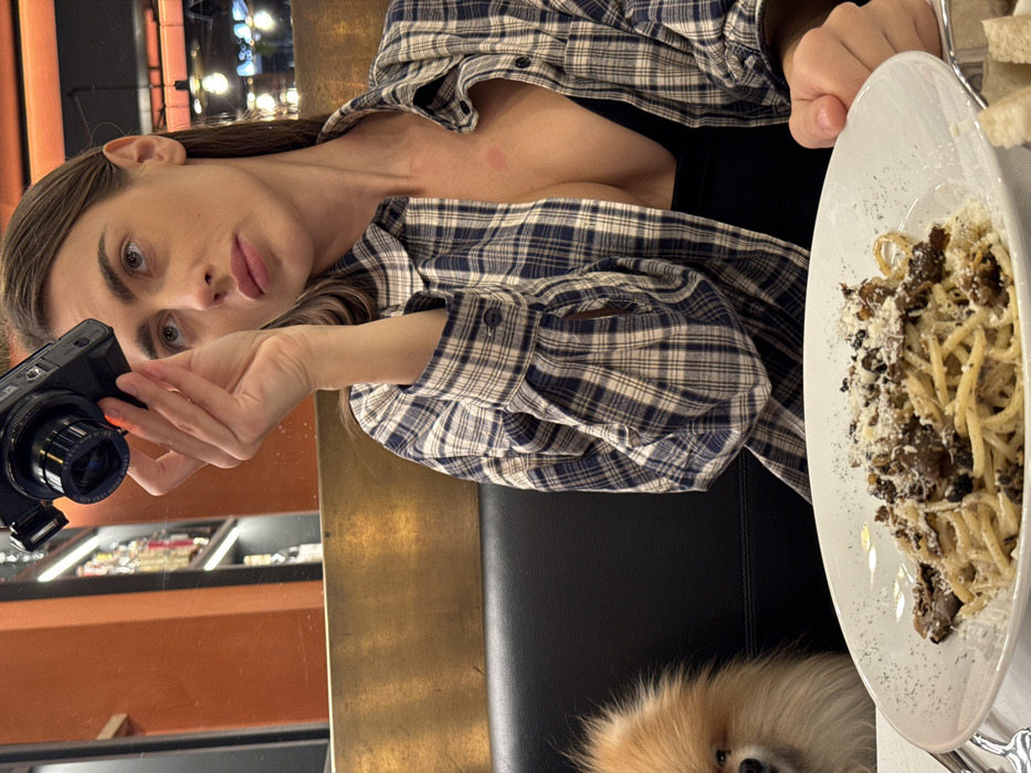 The image displays an indoor restaurant setting. A woman with long brown hair, wearing a blue and white plaid shirt unbuttoned over a black top, occupies a dark upholstered booth seat on the right. A reddish mark is visible on her upper chest. She holds a black compact camera in her raised right hand, pointed forward and slightly upward, while her gaze is directed similarly. A white plate containing spaghetti topped with black truffle shavings and grated cheese is positioned centrally on a white tablecloth. To the left of the woman, partially visible on the booth seat, is a small, fluffy, light brown dog, likely a Pomeranian. The background features a brass-colored divider, and reflections in a mirror show an orange wall and blurred shelves with bottles, suggesting a bar area. Overhead light fixtures are also visible. The specific location within Verona, Italy, cannot be determined from the visual information provided.