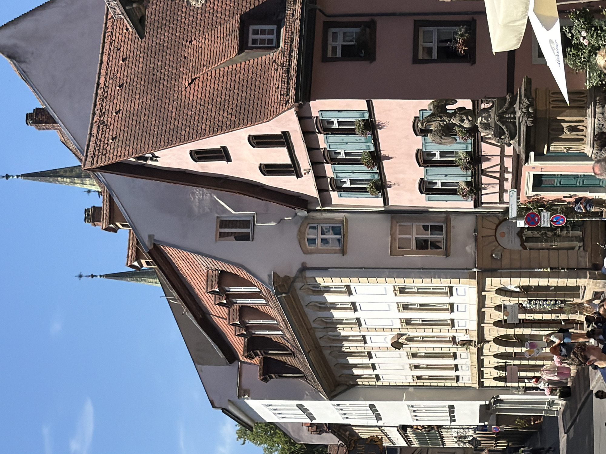 The image depicts a street scene in Bamberg, Germany, likely in the historic center given the architecture. In the foreground, people are visible walking and milling about on a cobblestone street. To the right, a cream-colored umbrella shades an outdoor seating area. Dominating the middle ground are several multi-story buildings with characteristic Bavarian tiled roofs and ornate facades. One building is painted a pale pink and features blue shutters on its windows. A stone fountain topped with a sculpture of an eagle stands prominently in front of this pink building. Other buildings are lighter in color, some with multiple arched windows and decorative stonework. In the background, church spires with crosses rise above the rooftops, indicating a religious presence in the vicinity. Trees are visible on the left side of the street, suggesting a blend of urban and natural elements. The sky is clear and blue, indicative of fair weather. The overall impression is that of a bustling, charming European city street on a sunny day.