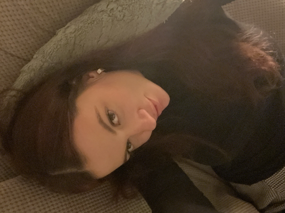 A woman with dark hair and fair skin rests on a textured gray surface, possibly a couch or cushion. She wears a black garment. Her head is tilted, her eyes are open, and she has a slight, subtle smile. She has a square-shaped earring in her left ear. The lighting is somewhat dim, creating a soft, intimate atmosphere.