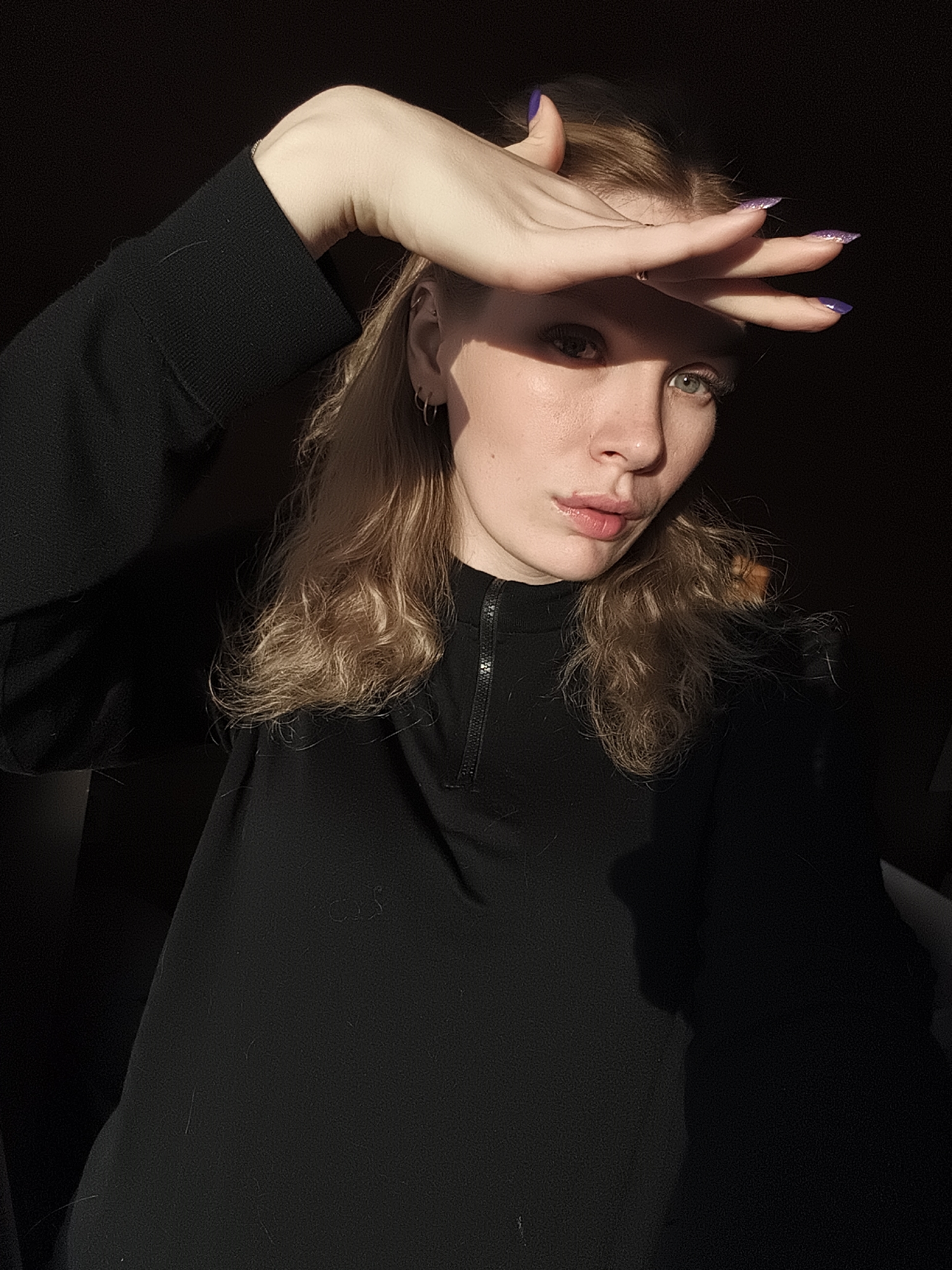A young woman is photographed indoors with dramatic lighting. She is fair-skinned, with light-colored hair and green eyes. Her left arm is raised, with her hand shielding her eyes from the sun. She wears a black top with a zipper. Her nails are painted purple. The background is a dark interior. The location is unclear.