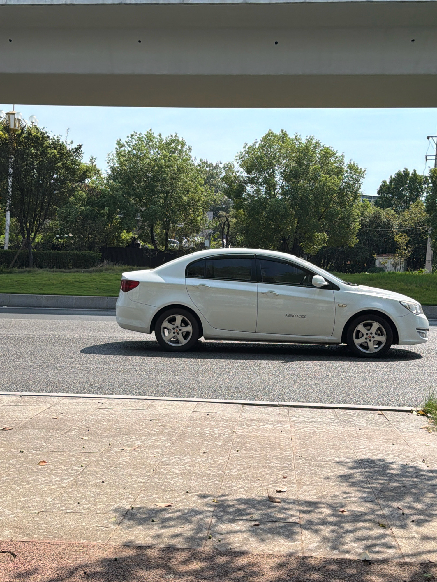 A white sedan with "AMINO ACIDS" printed on its side is driving on a paved road. The car is positioned in the center of the frame, moving from left to right. The road is bordered by a grassy median and trees. The sky is blue with some clouds. In the foreground, there is a concrete sidewalk with scattered leaves and shadows from trees. The scene appears to be in a city setting, possibly Jiangjin, China, given the architectural style and vegetation. There are no people visible in the image.