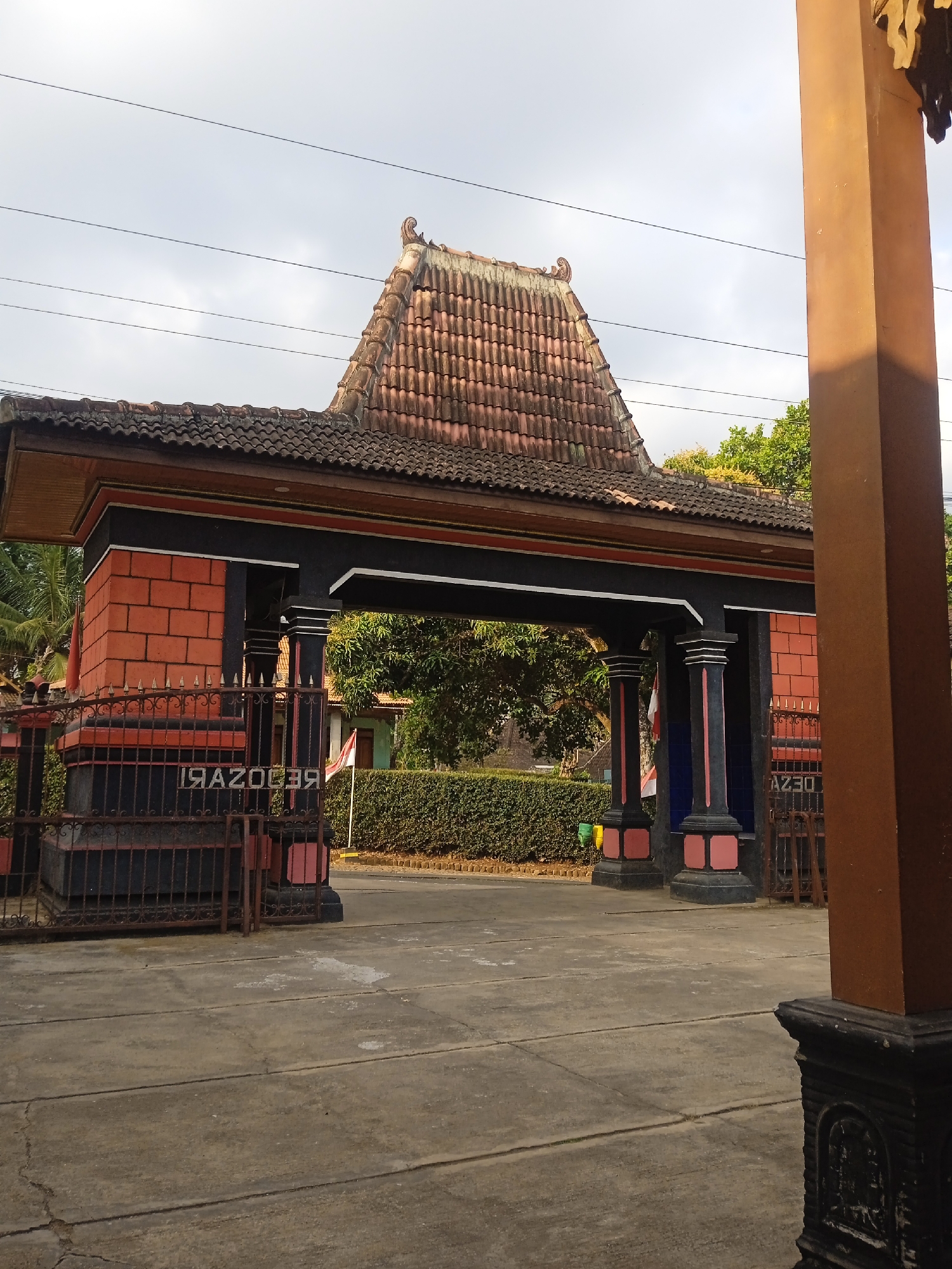 This image depicts a traditional Indonesian gate with a tiled, multi-tiered roof, set against a cloudy sky. The gate features prominent pillars painted black and pink, with brick-patterned walls on either side. A black metal fence with pointed finials encloses the area in front of the gate. Behind the gate, a neatly trimmed green hedge is visible, with trees and a building in the distance. A small Indonesian flag is also present. The ground in the foreground is paved with concrete slabs. The overall scene is static, with no people or animals visible. The gate appears to be an entrance to a property or establishment in the city of Sugihan, Indonesia, characterized by its distinctive architectural style.