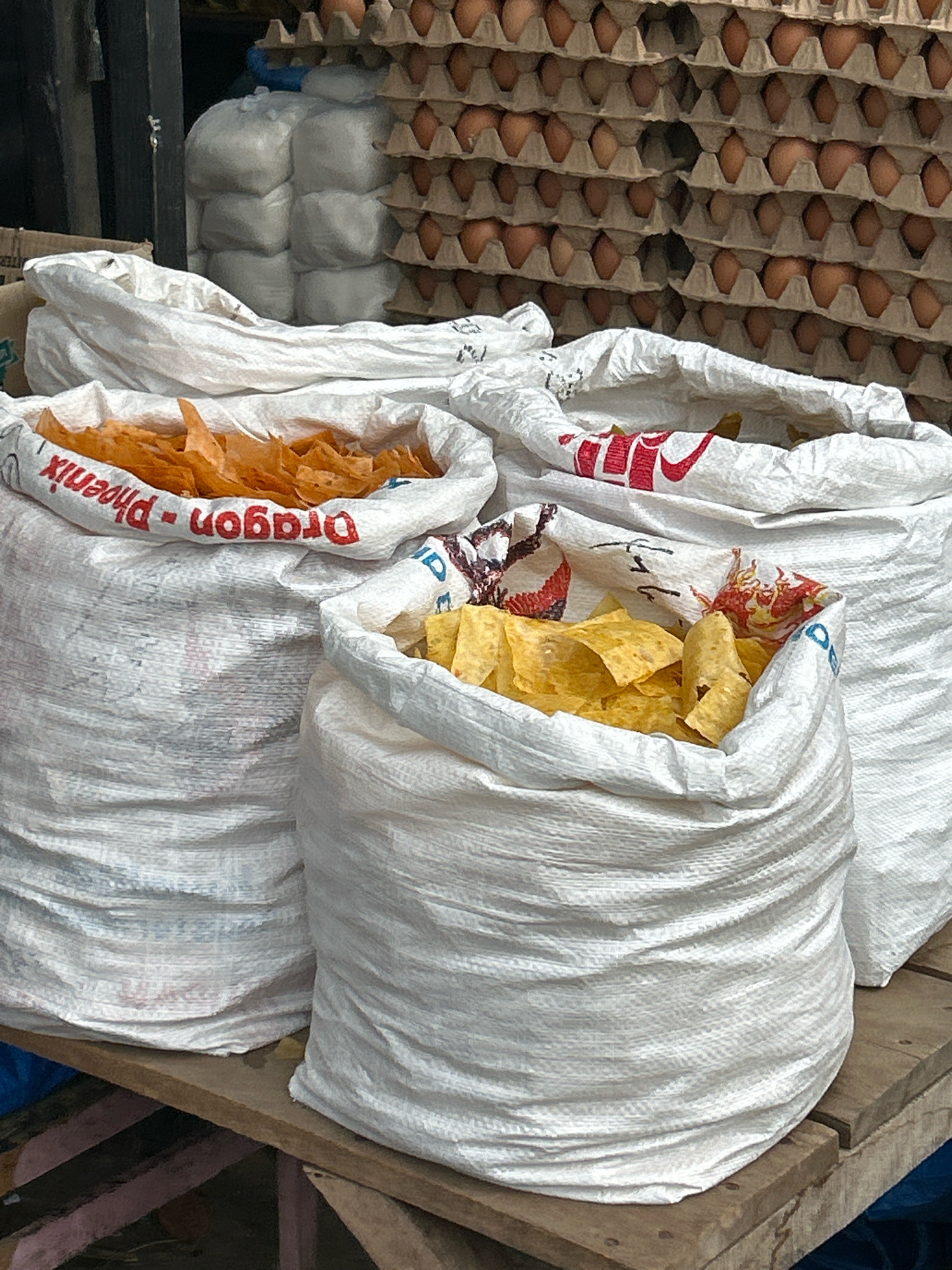 The image captures a close-up view of several large, white woven sacks filled with snacks, positioned on wooden planks. In the foreground, three sacks are visible. The leftmost sack, partially obscured, contains a pile of orange, triangular snacks, likely chips. The middle sack is overflowing with yellow, triangular snacks, also appearing to be chips. The rightmost sack, also partially filled, shows a glimpse of yellow chips. The sacks have printed text; the one on the left has red text that reads "Dragon-Phoenix". In the background, stacked egg cartons filled with brown eggs create a textured wall. To the left of the egg cartons, there are several white, opaque plastic sacks. The overall scene suggests a market or storage area in Banda Aceh, Indonesia, where food items are being prepared or stored for sale.