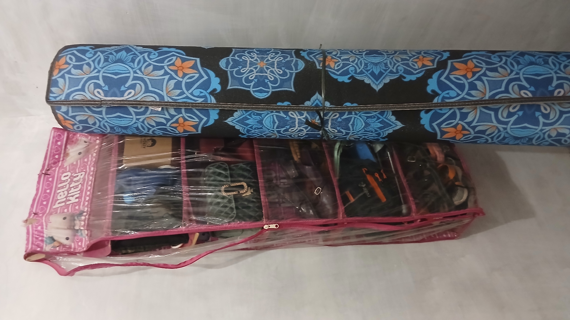 A vertically oriented, medium-shot, indoor image depicts an organizer bag and a rolled-up mat, likely in a storage area. The organizer bag, pink with a clear plastic front and a Hello Kitty graphic, hangs on the left side of the frame. It is filled with various items, including what appear to be purses, shoes, and boxes. The rolled-up mat, positioned to the right of the organizer bag, is black with an intricate blue and orange floral pattern. Both items are leaning against a light-colored, textured wall, possibly concrete. The lighting is even, suggesting an ambient indoor environment. No people are present in the image, and no specific actions or interactions are taking place. The image does not provide any discernible information about the city of Klambir Lima Kebun, Indonesia.