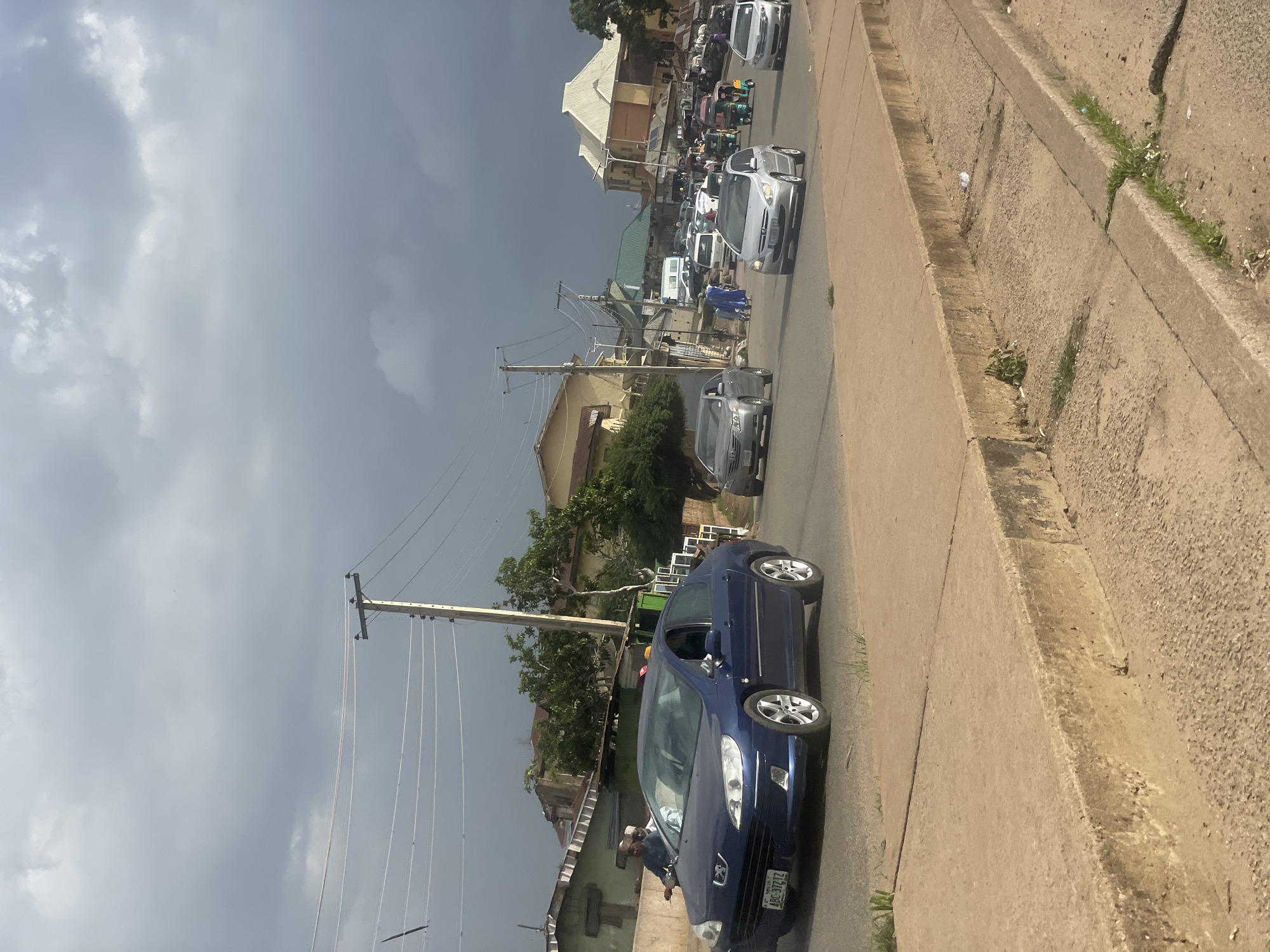 A medium shot captures a street scene in Jos, Nigeria, under a cloudy sky. A blue Peugeot sedan, license plate "ABC 3721Z", is in the foreground on the left side of the road. The car is in motion, and its driver is not visible. Behind the Peugeot, several other vehicles, including silver sedans, a white van, and a few tricycles, are proceeding down the paved road. The road is lined with buildings, some appearing residential and others commercial, with visible power lines and poles. The sidewalk in the immediate foreground is made of concrete and shows signs of wear and grass growing through cracks. People can be seen walking and riding a motorcycle on the left side of the road. The overall atmosphere suggests a busy urban environment.