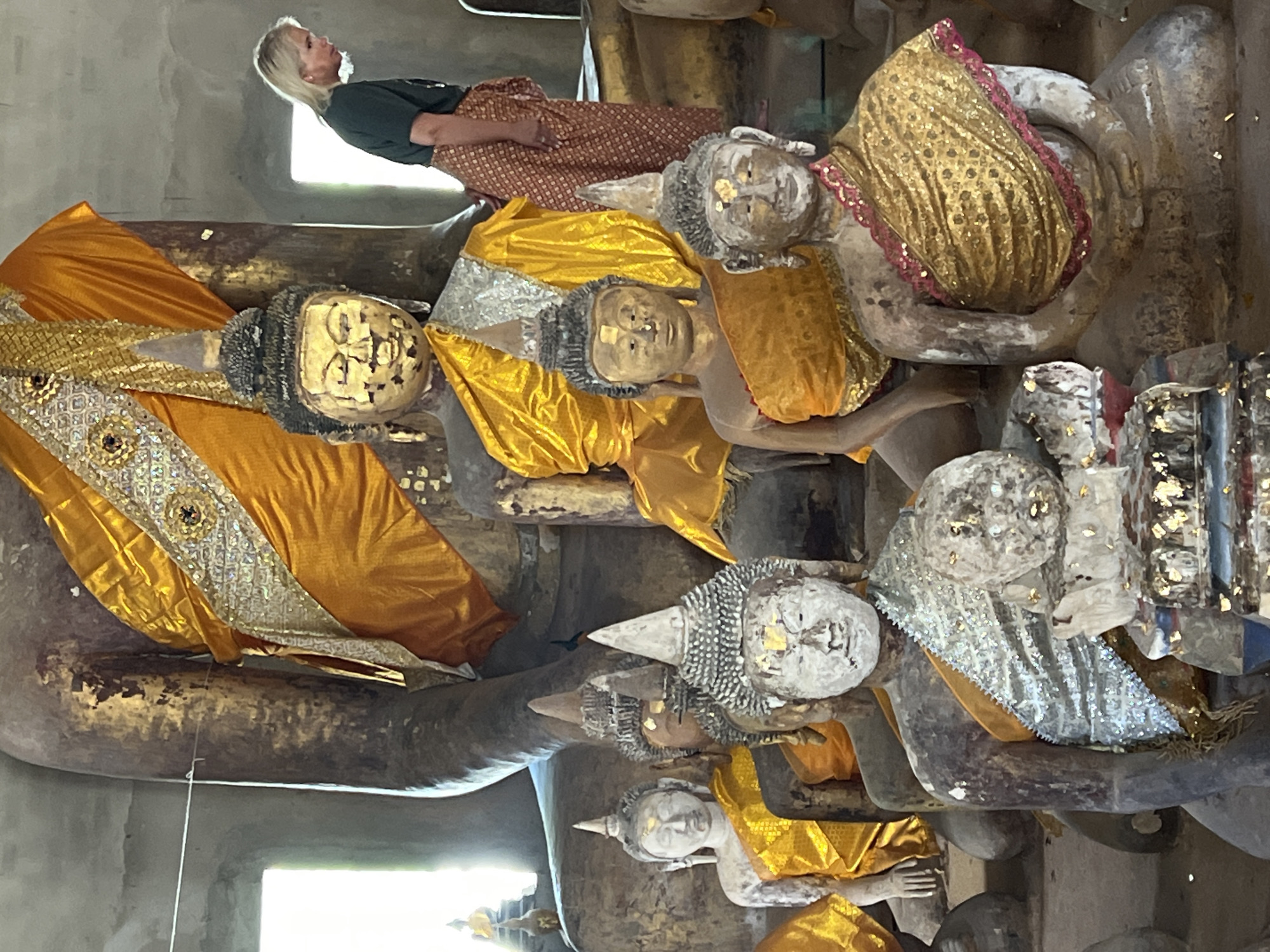 The image shows a collection of Buddha statues in a temple setting in Baan Harn, Thailand. A woman with blonde hair, wearing a dark green t-shirt and a patterned skirt, stands to the right, looking down. The foreground is dominated by several smaller Buddha statues, some with gilded faces and robes, others with weathered, stone-like appearances. They are adorned with yellow and silver fabric. Behind these, a larger, partially gilded Buddha statue is visible, draped in orange and gold cloth. The environment appears to be an indoor space with a concrete floor and textured walls, suggesting a historical or religious site. Natural light enters from windows on the left side of the frame.
