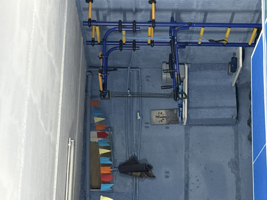 A cluttered indoor space exhibits a blue-painted wall. Hanging near the ceiling is blue and yellow gymnastic equipment. A multicolored triangular banner cascades down the wall alongside pipes. A worn, gray towel hangs beneath a wall-mounted clock. Other fixtures include a platform and a mounted speaker. The space's arrangement gives off an impression of storage or repurposed use.