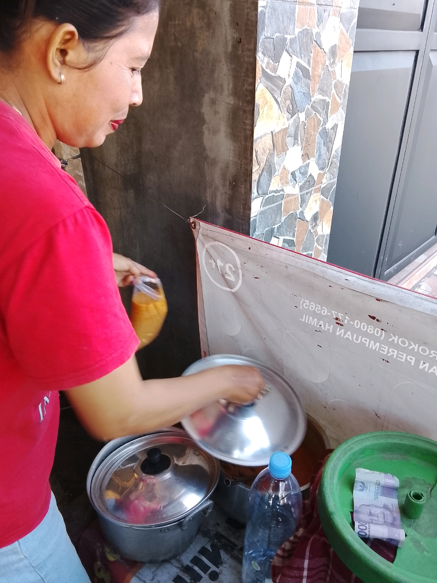 A woman in a red shirt is standing and preparing food. She is holding a clear plastic bag with orange liquid. In front of her is a large metal pot with a lid, and to the right is a green plastic bowl containing two folded Indonesian banknotes, a plastic bottle of water, and a roll of material. The background shows a concrete wall and a wall covered in colorful stone tiles. A white banner with text is draped on the wall. The scene appears to be outdoors, possibly a street food stall, in Damarsi, Indonesia.
