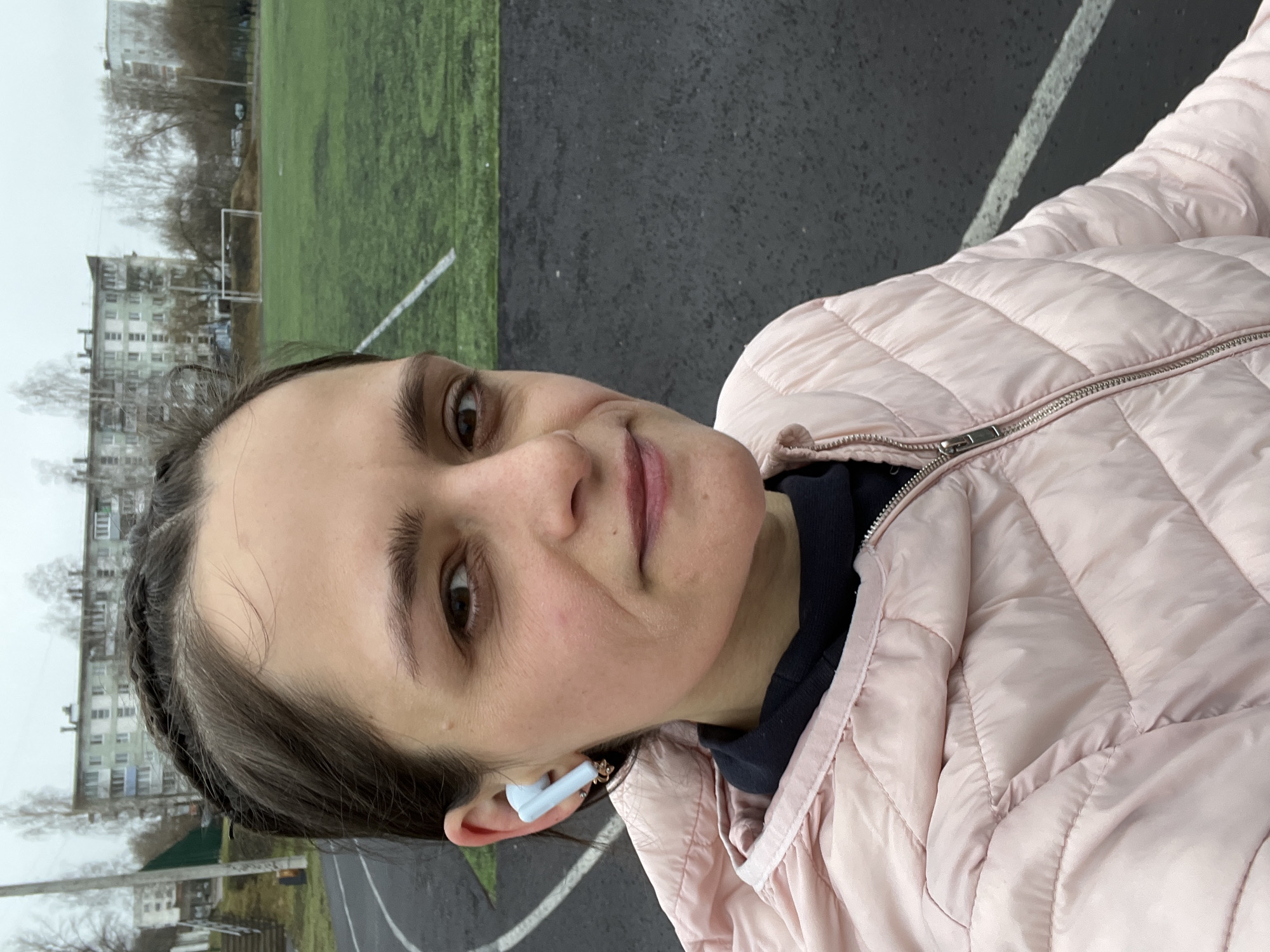 A woman wearing a light pink quilted jacket and white wireless earbuds is posing for a selfie. She has brown hair braided back from her face and brown eyes. Behind her is a sports field with green grass and a black running track. In the distance, buildings are visible, suggesting an urban environment in Chaikovsky, Russia. The sky appears overcast.