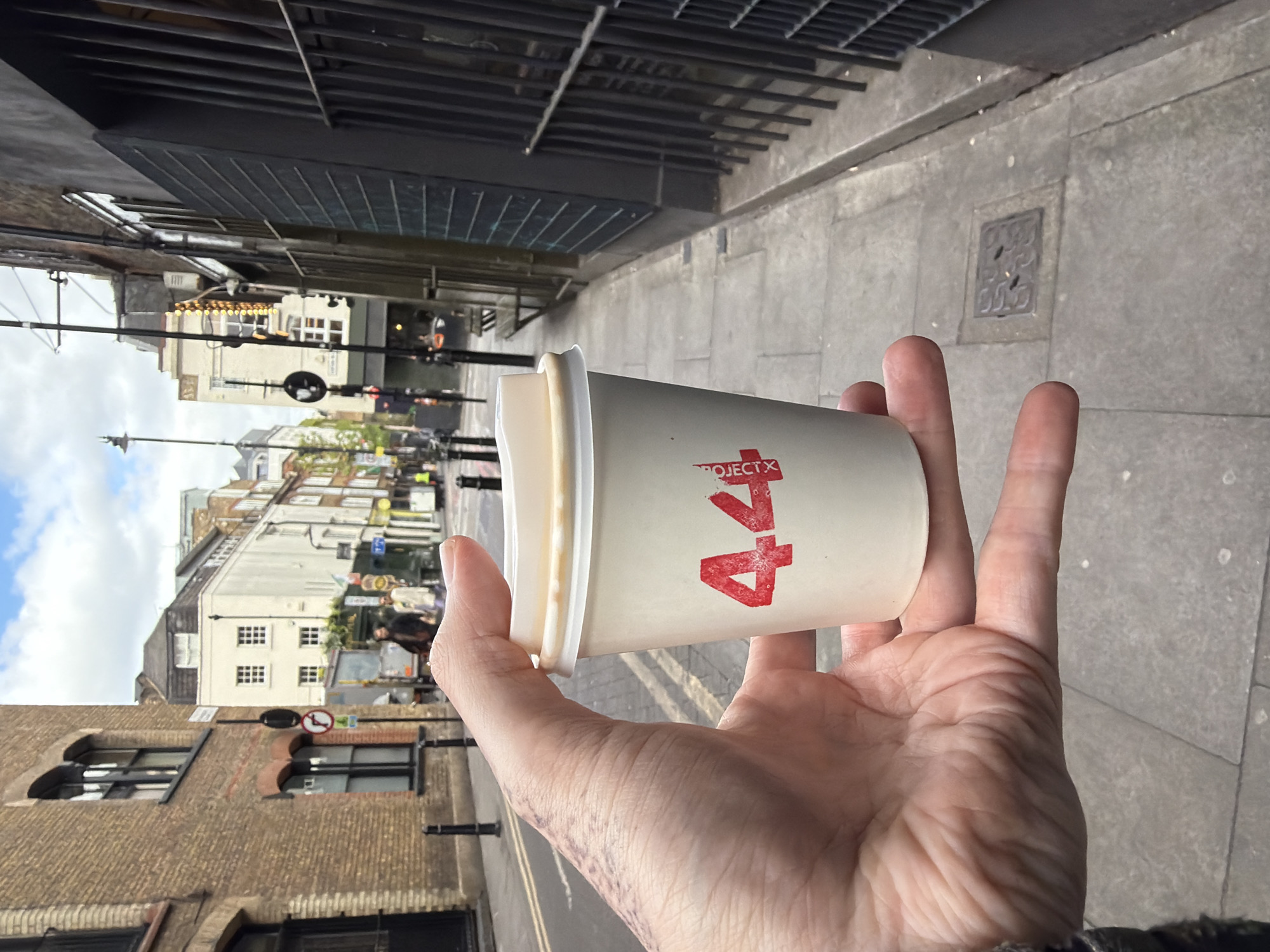 A medium shot shows a hand holding a white "Project 44" coffee cup, with a city street in the background. The street includes buildings of varying heights and architectural styles, ranging from brick to painted facades. Street signs, traffic lights, and pedestrians are visible. A metal grate structure appears above the hand on the left, while pavement and a manhole cover are on the right. The location is a street in London, United Kingdom.