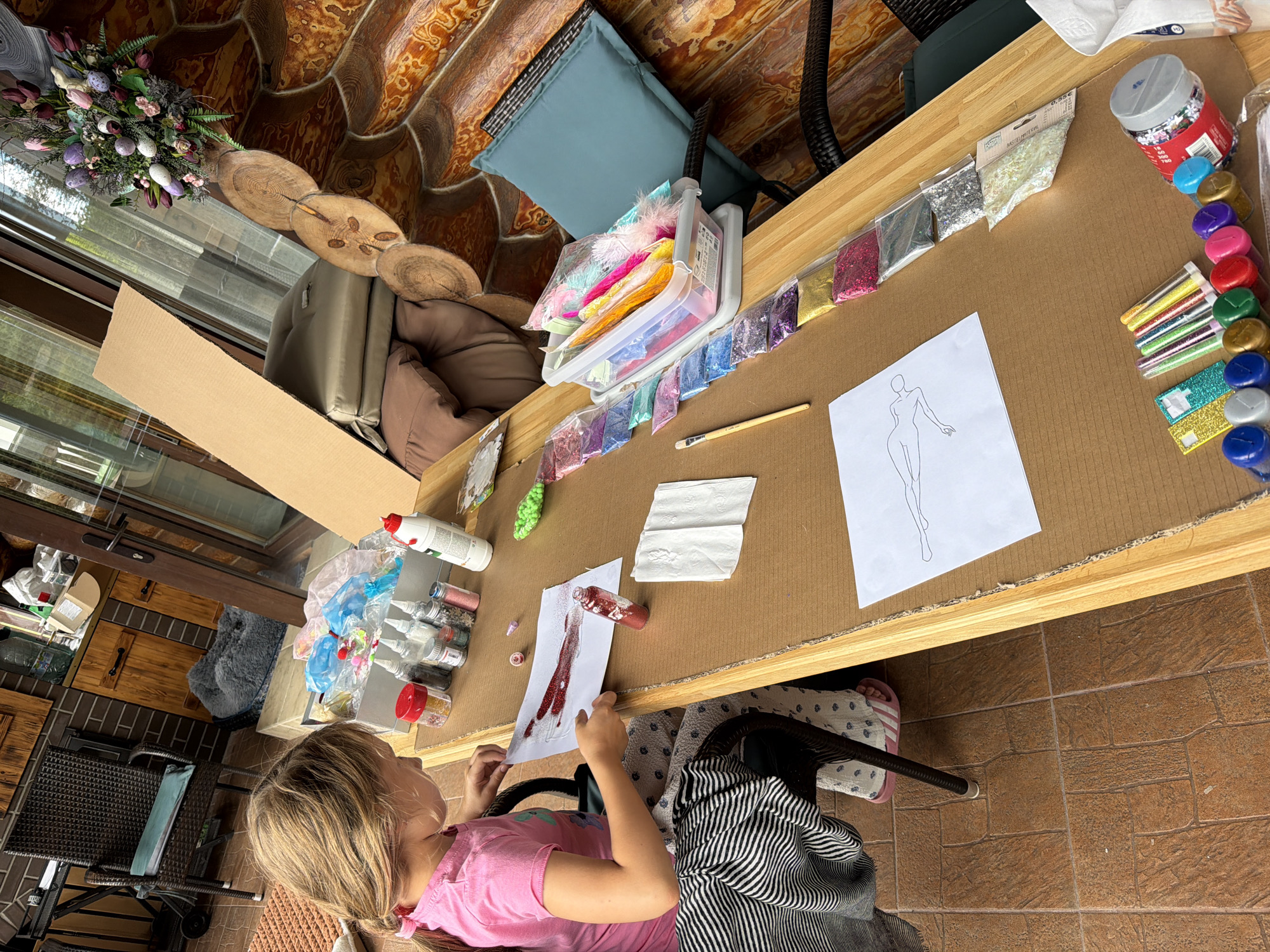 In Baloži, Latvia, a young girl with blonde hair in a pink t-shirt sits at a wooden table covered with brown cardboard. She is actively engaged in an art project, applying red glitter to a sheet of paper. Various craft supplies are spread across the table, including bottles of glitter, a container filled with colorful feathers, and a pencil. In the background, a wooden cabin structure is visible, with a patio area featuring chairs and plants.