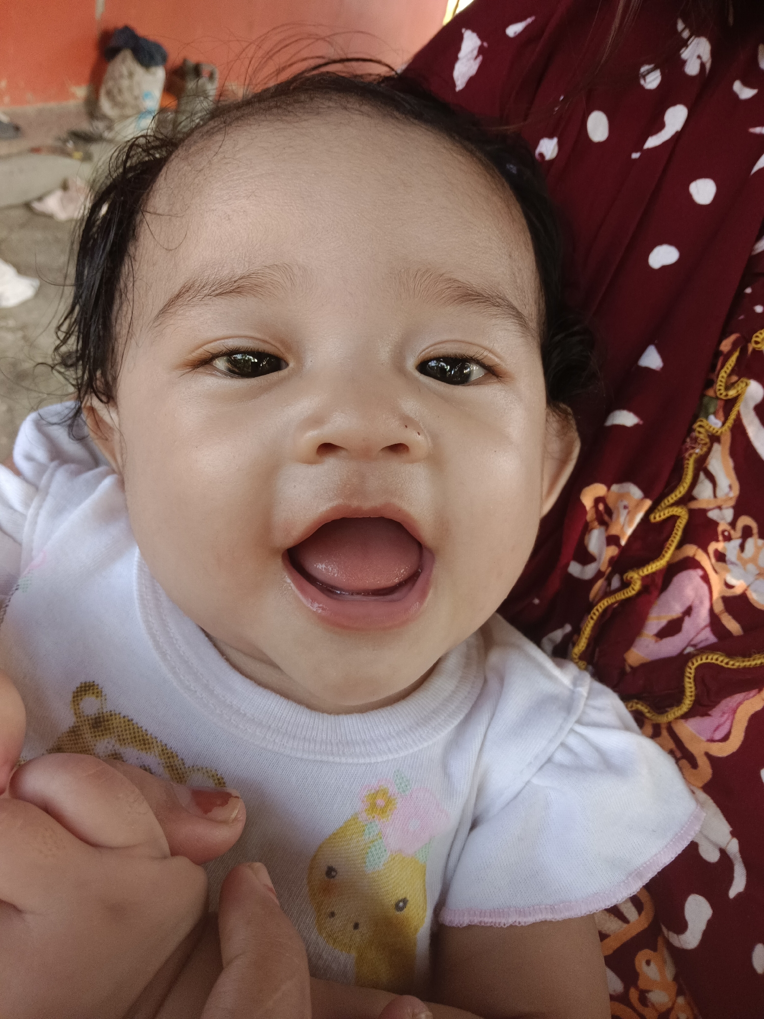 The image shows a close-up, overhead shot of a baby's face, looking directly at the camera. The baby has dark hair, brown eyes, and an open mouth. They are wearing a white onesie with a cartoon bear graphic. To the right of the baby, a person's arm is visible, draped in a dark red cloth with a white polka dot pattern. The background is out of focus and appears to be an outdoor environment, with hints of orange walls and what might be miscellaneous objects. The image was taken in Kawua, Indonesia.