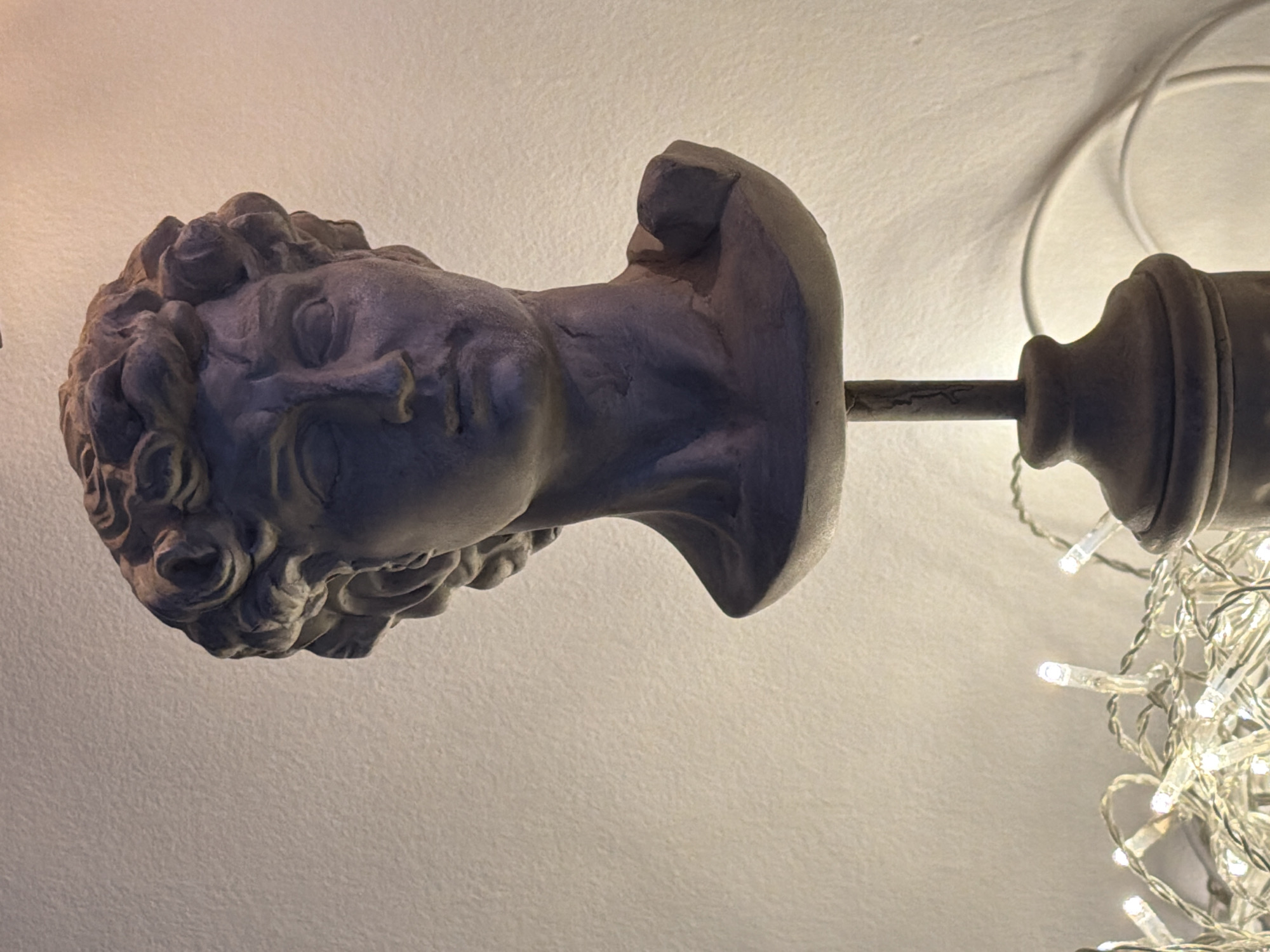 The image shows a close-up, horizontal shot of a grey, plaster bust of a classical head, possibly a Greek or Roman sculpture. The bust is positioned on a metal rod that extends to the right, where it connects to a larger, decorative finial. This finial is adorned with a string of small, white LED lights, which are illuminated and create a festive atmosphere. The background is a plain, textured off-white surface, likely a wall or ceiling. The lighting suggests an indoor setting, possibly at night or in a dimly lit room, as the string lights are the primary light source in the immediate vicinity. The bust itself has detailed curly hair and facial features, with the eyes closed and a serene expression. The overall composition, with the classical bust combined with modern decorative lights, creates an interesting juxtaposition of old and new. There is no visible text in the image. The context of Tel-Aviv, Israel, does not provide specific visual cues within the image itself, but suggests the cultural and geographical setting.