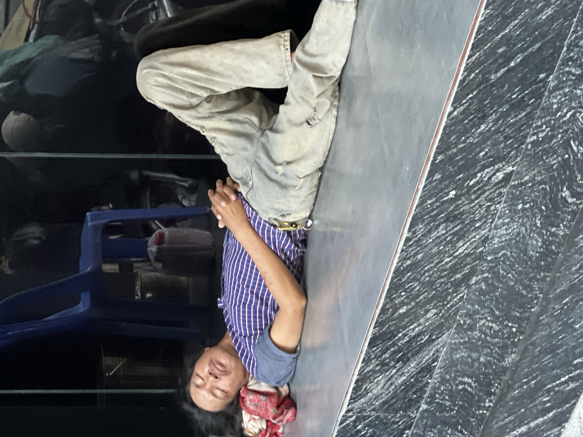A man is sleeping on a grey surface, which appears to be a step or ledge, in front of a dark background. He is lying on his back with his head resting on a red and white patterned cloth. He is wearing a blue and white striped t-shirt and light-colored, distressed pants. His eyes are closed, and he has a mustache. To his left, a blue plastic chair is visible, and behind him, various items including what appear to be buckets and bags are present. The scene is dimly lit, suggesting it might be outdoors in a shaded area or indoors with limited lighting. The surface he is sleeping on has a marble-like pattern. The overall impression is of a person taking a rest in a public or semi-public space.