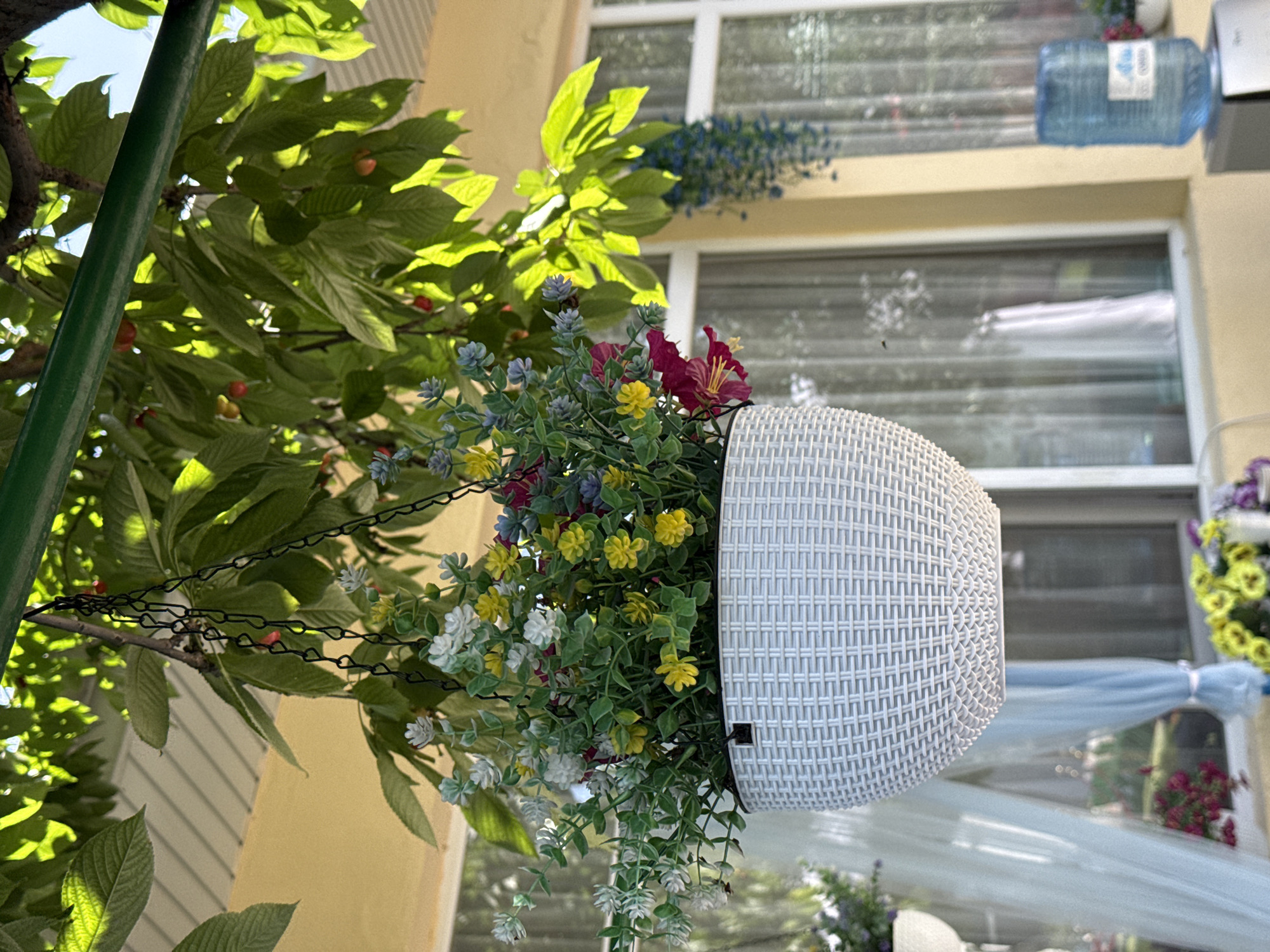 The image shows a hanging flower basket with a white woven container, filled with various colorful artificial flowers including red, yellow, white, and purple hues. The basket hangs by a black chain attached to a tree branch with green leaves, partially obscuring the left side of the frame. In the background, a light yellow building facade is visible, featuring windows with white frames. A water bottle is attached to a window on the upper right. Beneath the hanging basket, there is a suggestion of an outdoor setting, possibly a patio or garden, with draped fabric and other floral arrangements. The location is described as Unknown, Unknown.