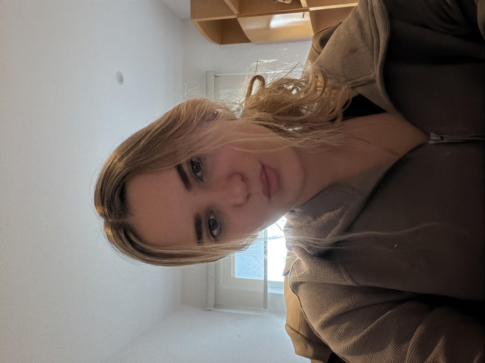 An image taken in an indoor setting, identified as San Bernardino Tlaxcalancingo, Mexico, depicts a young adult female from the chest up, centered and facing the camera directly. She has light-colored hair, partially pulled back into a loose ponytail, with strands falling forward. Her facial expression includes slightly puckered lips in a pout. She is wearing a dark, possibly olive-green or brown, zippered jacket with a textured fabric visible on the shoulders and a dark garment underneath. A thin necklace is visible around her neck. The background to the subject's left features a white wall with a window revealing a bright exterior, possibly a blue sky. To the subject's right, a light brown wooden shelving unit with curved elements is partially visible. The room has white walls and ceiling, with a recessed light fixture discernible on the ceiling.