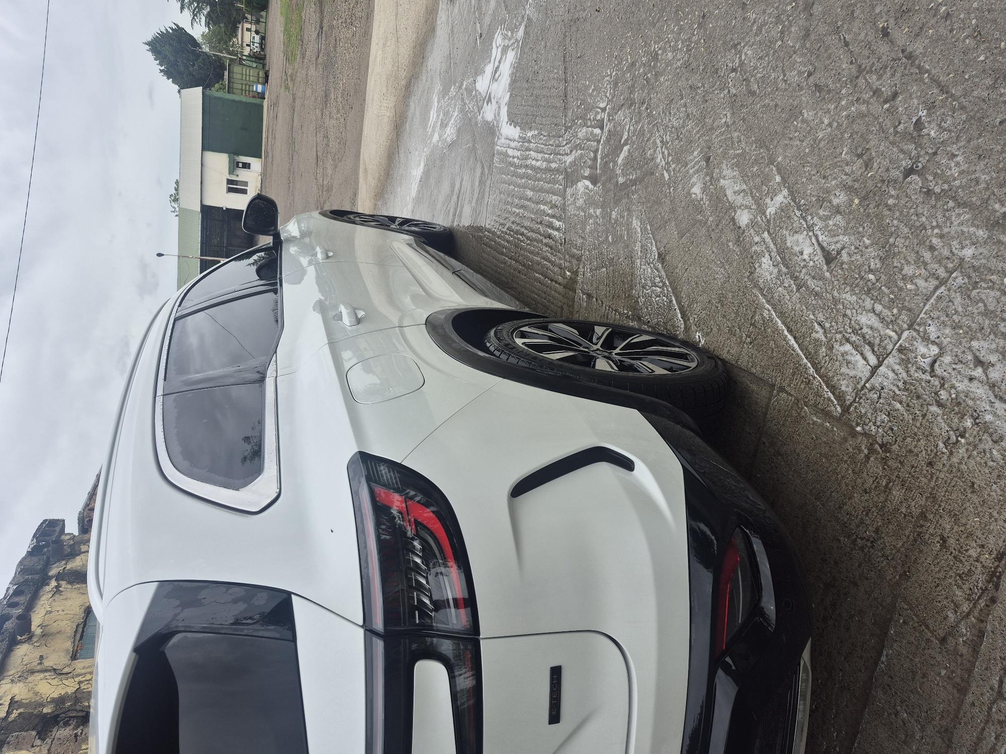 A white Peugeot SUV is parked on a concrete surface with soap suds visible, indicating a recent wash. The rear of the vehicle is in focus, showing the taillights, bumper, and the "E-TECH" badge. To the left of the car, a multi-story building with aging facade is visible. In the background, a green warehouse-style building is present. Above it, some trees and a cloudy sky are visible. The photo was taken in Tbilisi, Georgia.