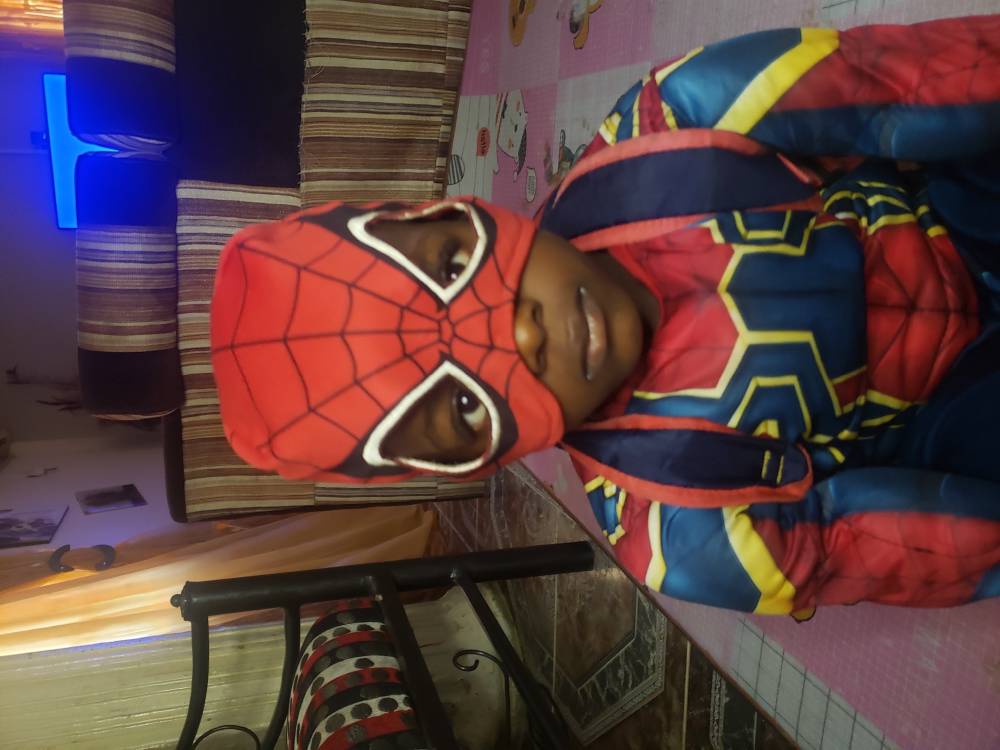 A young child, dressed in a Spiderman costume, is the sole subject of the image. The child is positioned in the center of the frame, facing forward with their head slightly tilted to the right. The Spiderman mask covers the child's face, revealing only their eyes and mouth. A blue and red backpack is visible over the child's shoulders, and the costume features intricate patterns in red, blue, and yellow.
The environment suggests an indoor setting, possibly a home. In the background, a sofa with striped upholstery is partially visible. To the left, a chair with a patterned cushion and a metal frame is present. A curtain, appearing to be made of sheer fabric and colored orange or yellow, hangs in the background, with a blue light source visible through it. The floor is covered with patterned tiles and a pink mat with cartoon graphics. The overall lighting is dim, with a noticeable blue glow emanating from the light source in the background. The image is a close-up, focusing primarily on the child and their costume.