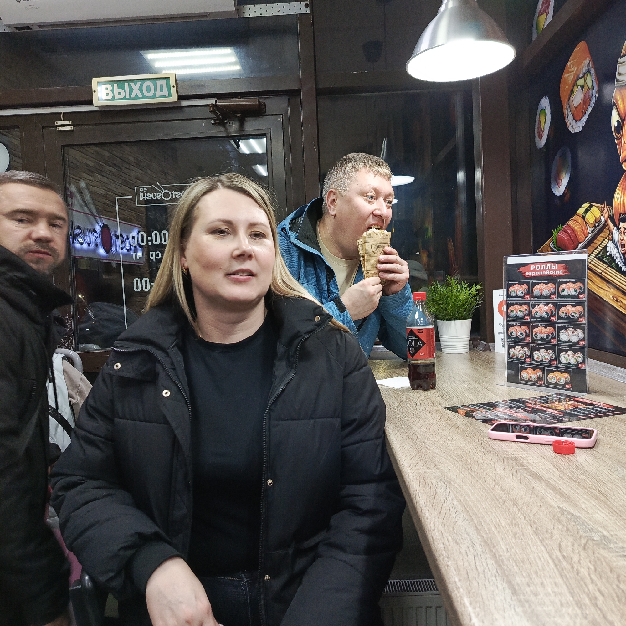The image captures a scene inside a sushi restaurant. A woman in a black puffer jacket and black shirt sits at a light wood counter. To her left, a man in a black jacket stands beside her. Behind them is a glass window with reflected writing. Further back, a man wearing a blue jacket is eating from a brown paper container. On the counter is a bottle of "Cola," a small potted plant, a sushi menu, a promotional menu, and a pink smartphone with a red object beside it. A silver light fixture hangs from the ceiling. The green "Exit" sign is above the door. The city name, Chaikovsky, Russia, can be inferred from the Russian text and the appearance of the environment.