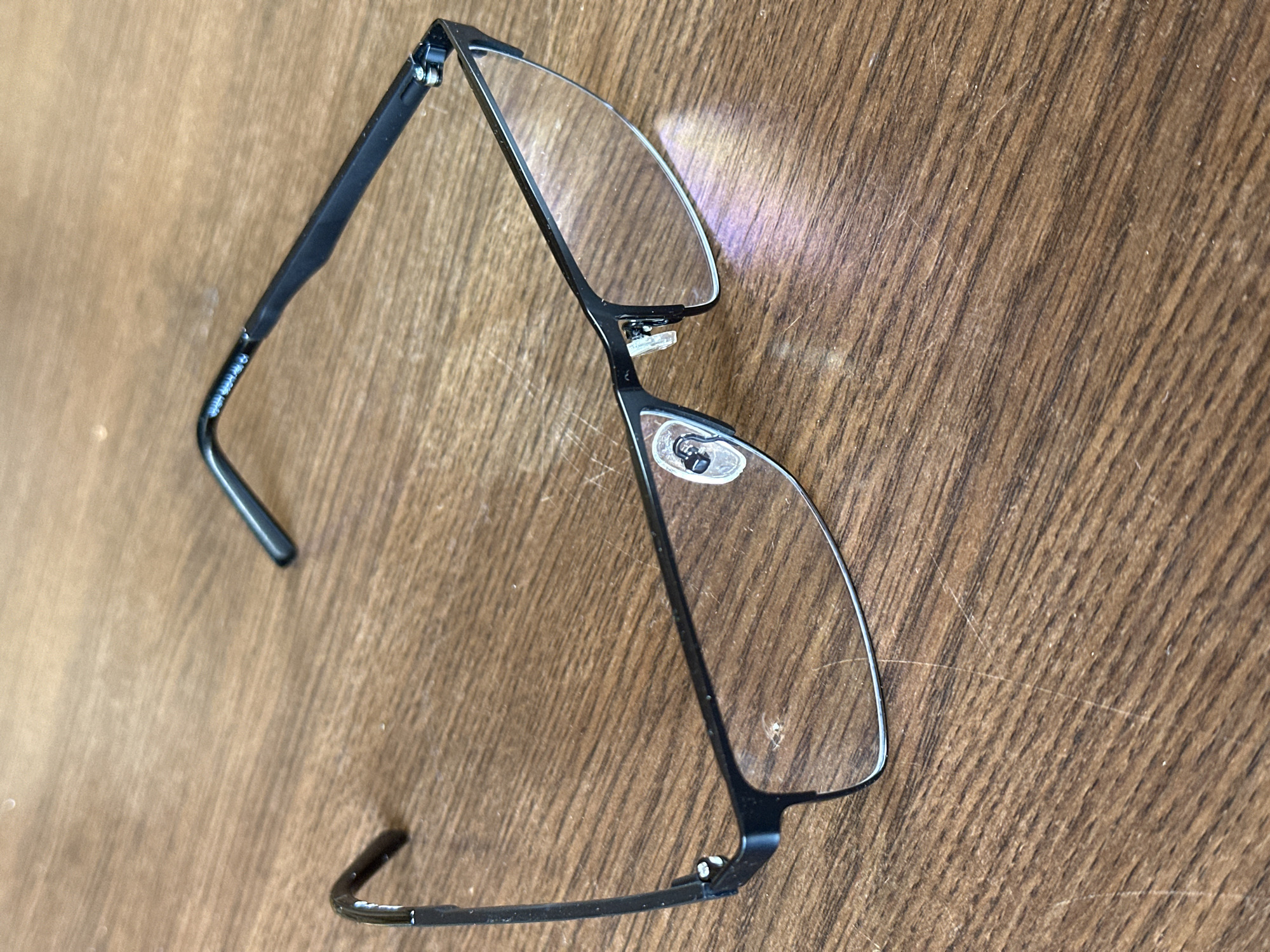 A pair of black-rimmed eyeglasses is situated on a wooden surface. The eyeglasses are rectangular in shape with thin black arms. The left arm appears to be slightly bent upwards. The lenses are clear and show some reflections. The wooden surface has a visible grain pattern and appears to be a tabletop or desk. There is no indication of the location in the city of Izobilnyy, Russia.