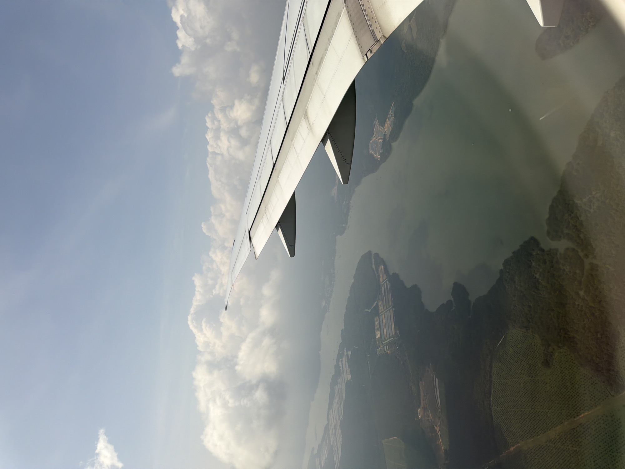 An aerial view from an airplane window shows a body of water with several boats on it, as well as land with lush green trees and some rectangular structures. The airplane's wing is visible in the foreground, with its white and gray surface taking up the right side of the image. In the sky, white, fluffy clouds are scattered against a light blue background. The scene depicts a landscape that appears to be a coastal area of Singapore, possibly Jurong Island, with its industrial complexes and waterways.