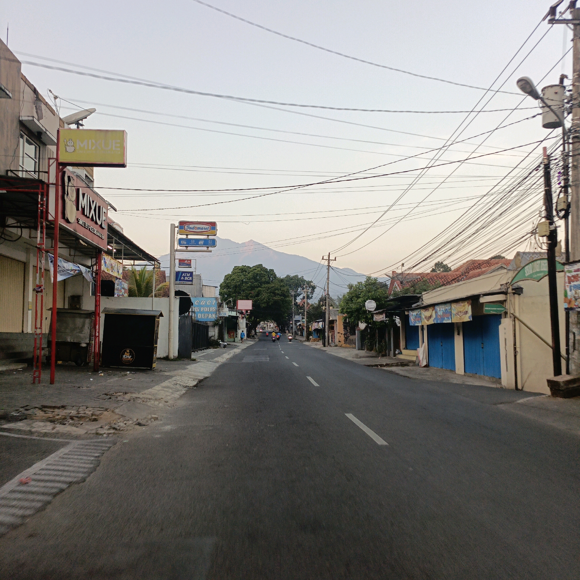 The image shows a street scene in Salatiga, Indonesia. The street is paved and has white lane markings. On the left side of the street, there are several shop fronts, including a "Mixue" ice cream and tea shop, with a red sign. Further down the street, there are more shops and residential buildings. On the right side of the street, there are also shops and residential buildings, some with blue shutters. In the background, there is a large tree and a mountain. The sky is overcast. There are several utility poles with many wires strung between them. A few motorcycles are visible on the street.