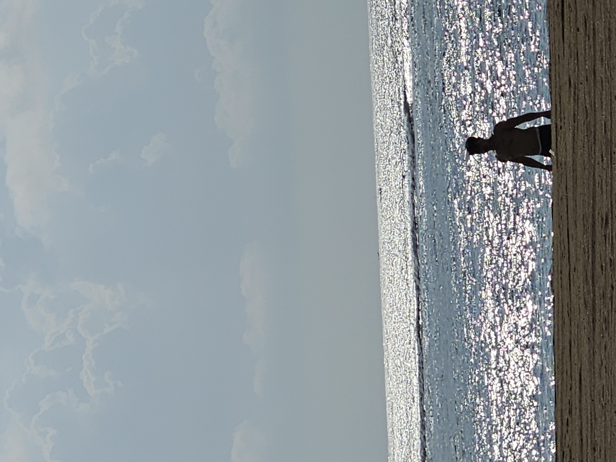 A lone, silhouetted man stands in shallow water at a beach in Denpasar, Indonesia. He is facing forward, with his arms at his sides. The water is clear and reflects the sunlight, creating a shimmering effect. The sky is pale blue with scattered white clouds. The beach is made of sand. The overall scene is serene and peaceful.