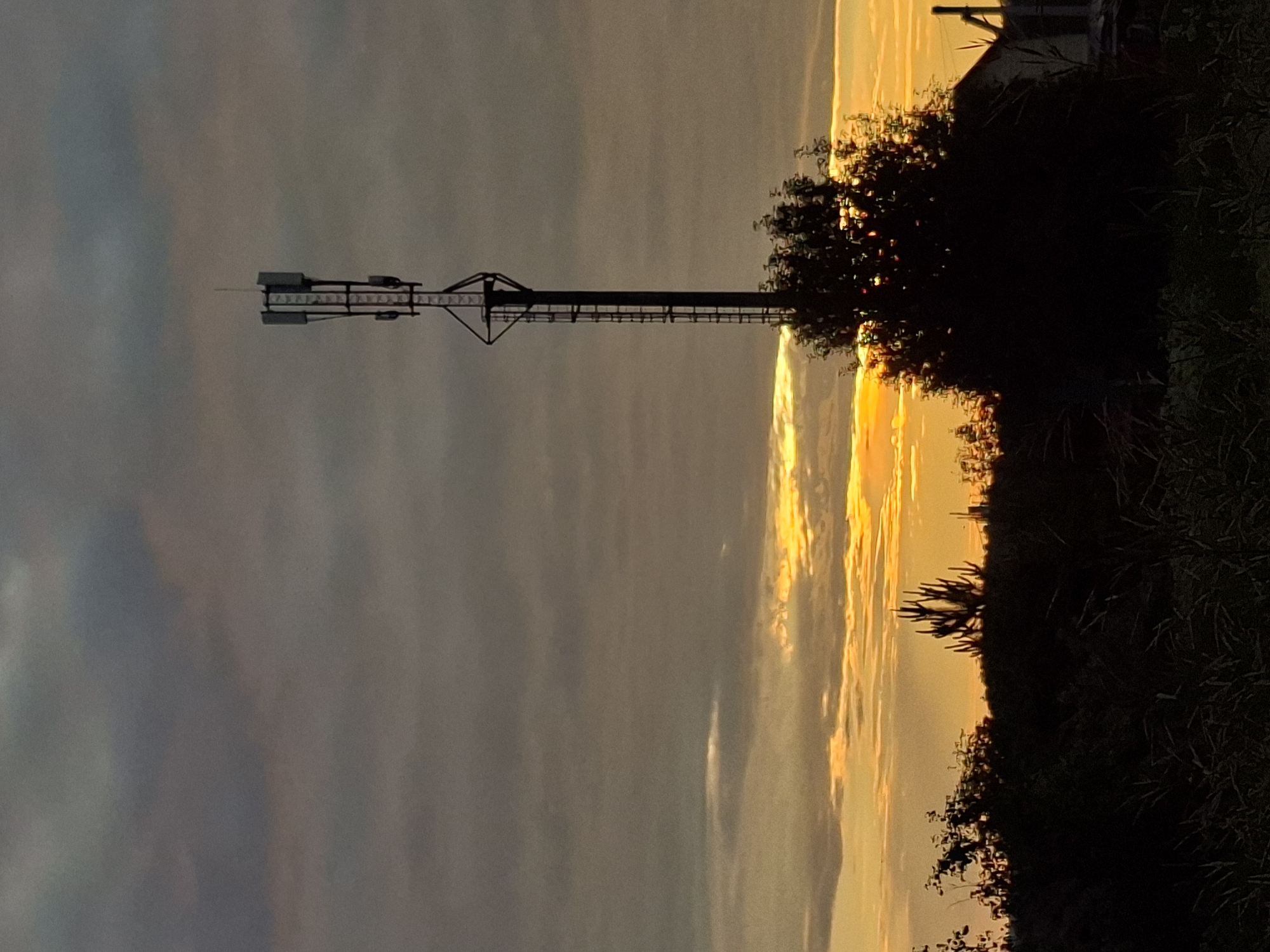 The image depicts a silhouette of a cell tower against a dusky sky. The sky is a mix of dark gray and orange clouds, indicating either sunrise or sunset. The cell tower is tall and slender, with several antennas mounted at the top. In the foreground, there is a silhouette of a tree and some vegetation. The overall scene is dark and moody, with the bright sky providing a stark contrast to the shadowed landscape. The location is identified as Unknown, Unknown.