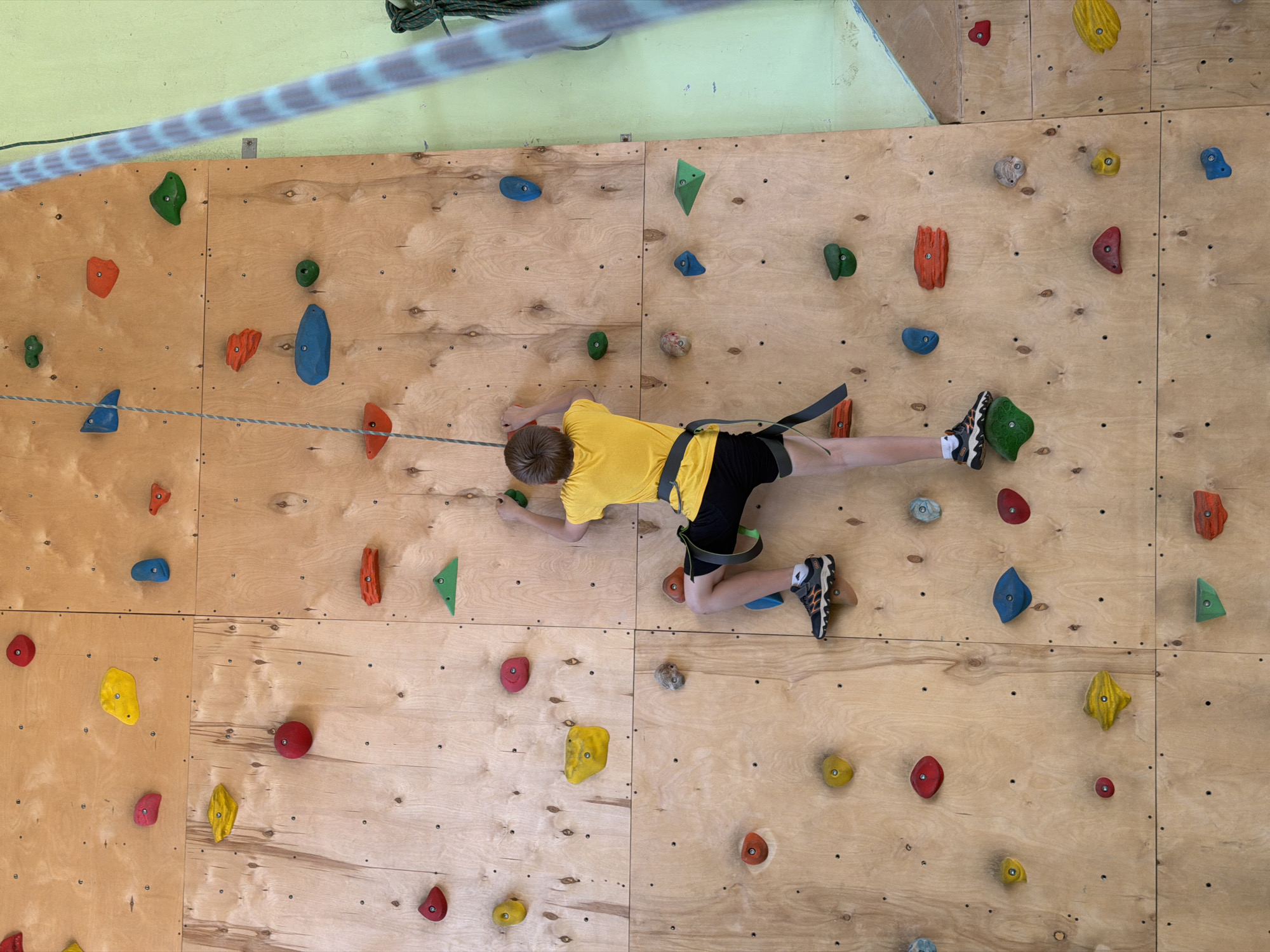 A person wearing a yellow shirt and black shorts is climbing a climbing wall. The wall is wooden with various colored climbing holds. The person is wearing a harness and is attached to a rope. The image was taken indoors. The location in the city of Izhevsk, Russia, is not explicitly mentioned or visible in the image.