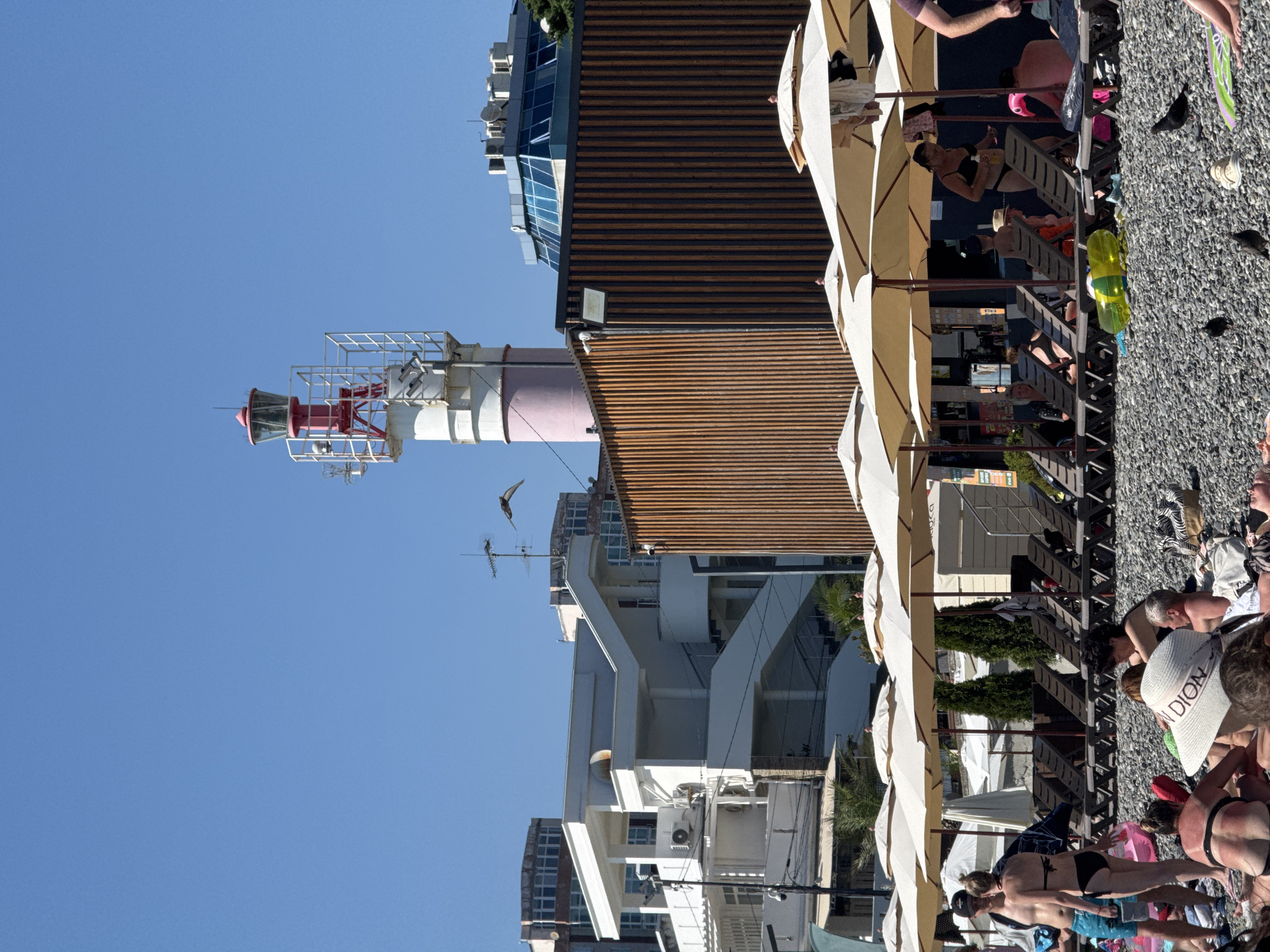 A bright, sunny day at a pebble beach in Sochi, Russia, features a prominent lighthouse with a red top and white body. Behind the lighthouse, a modern building with a blue glass roof is visible, along with other multi-story structures. In the foreground, numerous beach umbrellas provide shade over rows of empty loungers. People are scattered across the beach, some sunbathing on towels, others sitting or standing. A few individuals are in swimwear, suggesting a relaxed beach atmosphere. The ground is covered in small pebbles, characteristic of many Black Sea beaches. Pigeons are present on the ground. The overall scene depicts a lively day at a popular tourist destination.
