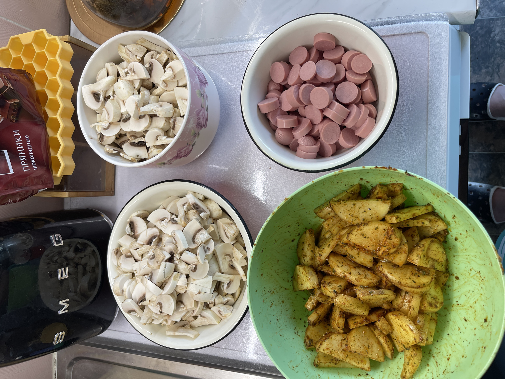 Four bowls of ingredients are arranged on a white countertop. In the upper left, a black SMEG toaster is partially visible. To its right, a large bowl contains sliced mushrooms. Below this bowl, a green bowl is filled with seasoned potato wedges. To the right of the green bowl, another bowl holds sliced hot dogs. In the background, a yellow honeycomb-shaped ice cube tray is visible, with a package of cookies labeled "ПРЯНИКИ шоколадные" (Gingerbread chocolate) placed on top of it. A dark, dome-shaped object, possibly a cake, is partially visible on the far right. The scene appears to be an indoor kitchen setting.