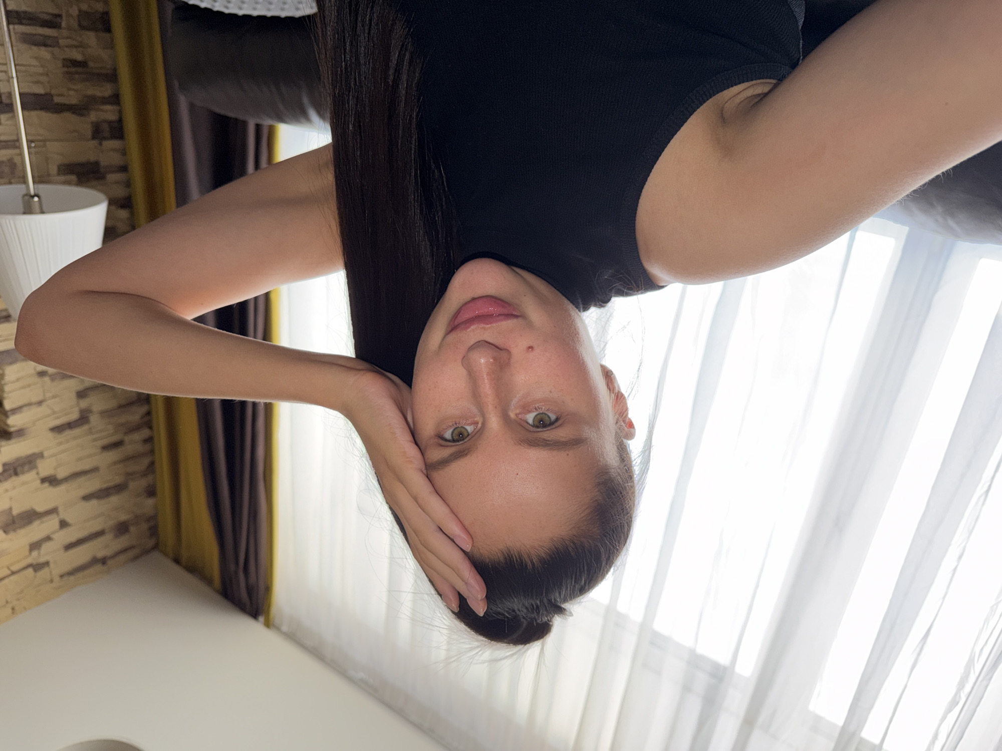 An inverted portrait features a young woman looking directly at the camera. She appears to be in her 20s or 30s, with fair skin, light brown/green eyes, and dark, long hair that is partially pulled back into a bun but with some strands falling forward over her left shoulder and arm. She is wearing a black sleeveless top. Her left hand is gently placed against the right side of her face, with her fingers resting on her forehead and temple. Her expression is neutral to slightly contemplative, with her lips slightly parted.
The setting is indoors, likely a room in a residence in Khimki, Russia, indicated by the location context. To the left of the woman, a wall with a textured, stone-like pattern is visible, along with part of a white modern lamp or light fixture. In the background, behind the woman, sections of gold and dark brown curtains frame a window. Bright natural light streams through sheer white curtains covering the window, suggesting it is daytime. A dark piece of furniture, possibly a sofa or headboard, is partially visible behind her head. The floor is light-colored and indistinct. There is no visible text in the image.