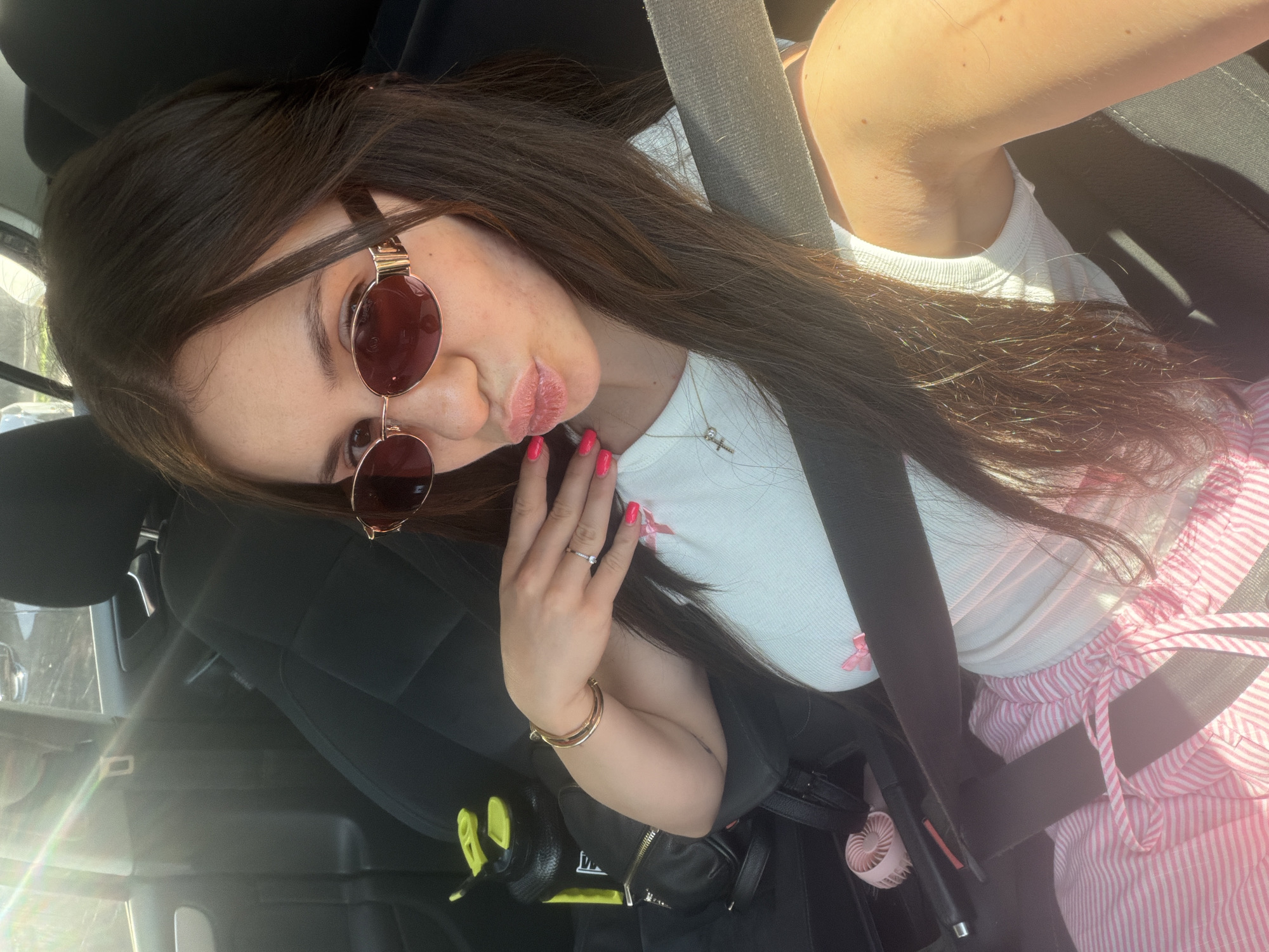 A woman is taking a selfie inside a vehicle. She is wearing sunglasses, a white top, and pink and white striped pants. The seatbelt is across her body. In the background, there is a car interior. The location cannot be determined to be Plovdiv, Bulgaria from the image provided.