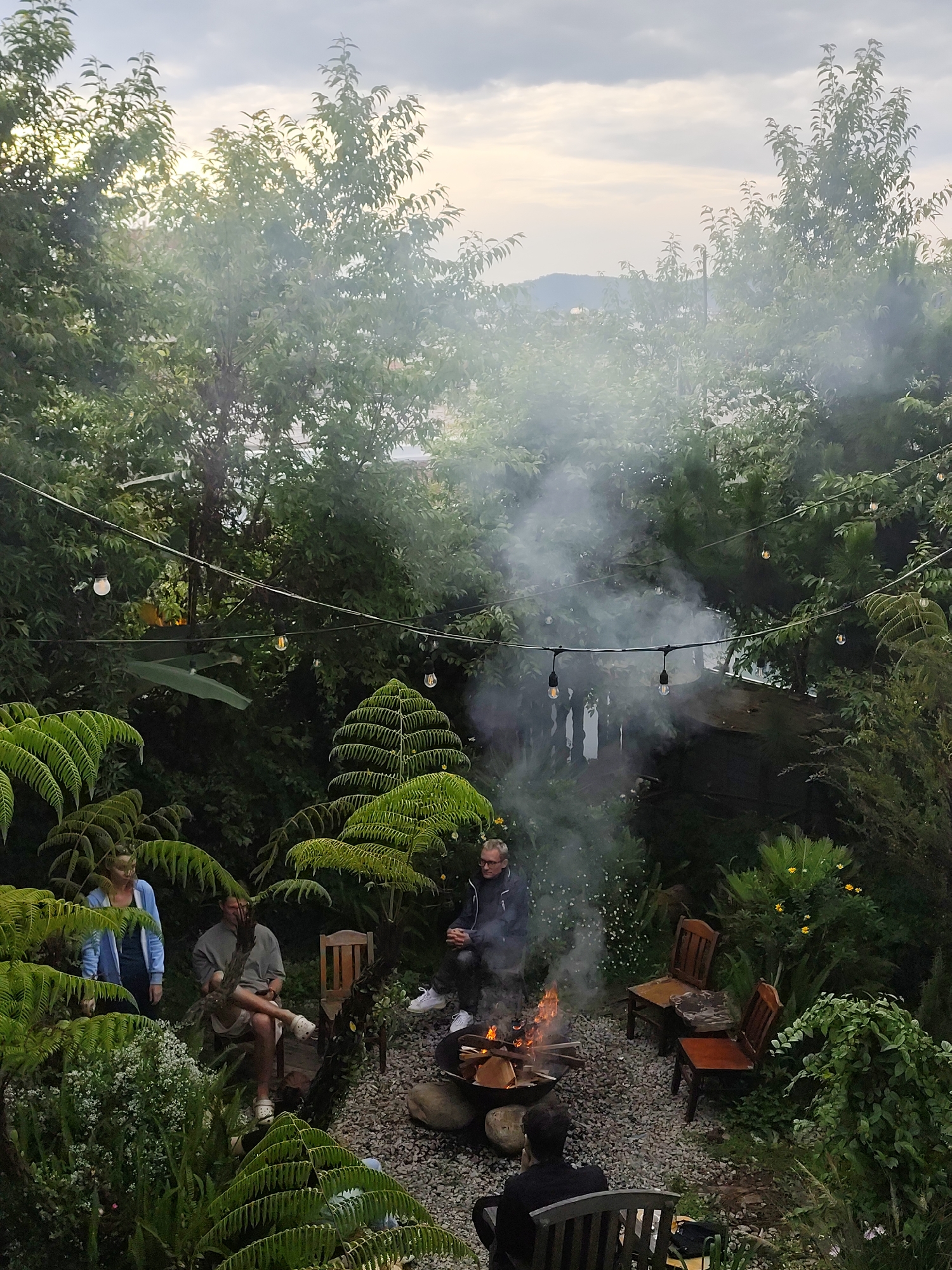 A group of people are gathered around a fire pit in a lush garden, which is potentially located in Lâm Viên - Đà Lạt, Vietnam. The garden is filled with ferns and other greenery. String lights are hung above the fire pit. There are five visible individuals. One person is tending the fire, while the others are seated in chairs surrounding the fire pit. The sky is overcast.