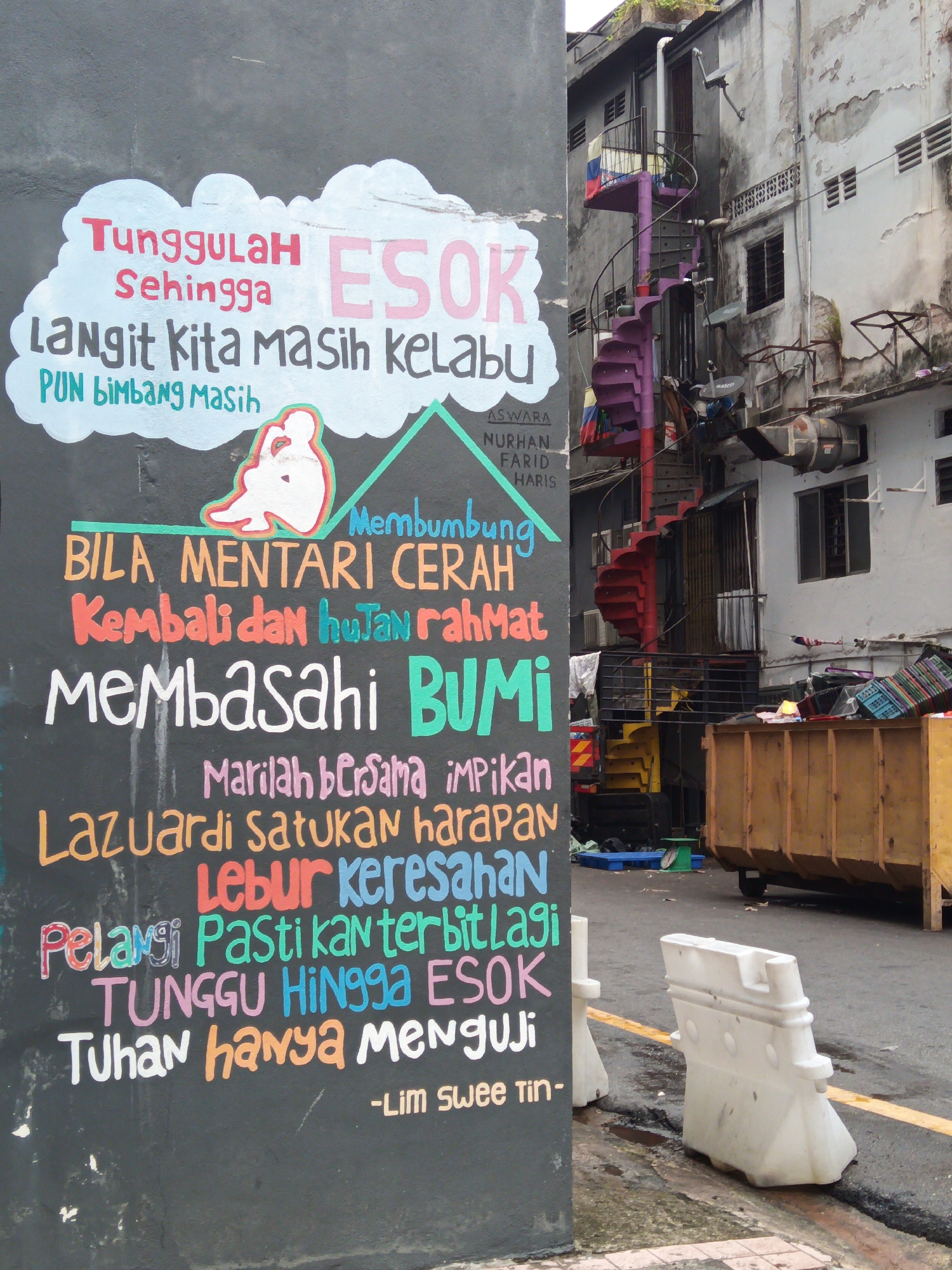 The image depicts a section of a street in Kuala Lumpur, Malaysia, characterized by a large, dark grey mural painted on a concrete wall. The mural contains a poem written in colorful, stylized lettering. Above the text, a light blue cloud graphic is painted, with the words "TUNGGGULAH Sehingga" in red and pink, and "ESOK" in large pink letters. Below this, "LANGIT KITA MASIH KELABU" is written in black. A small, simplified drawing of a person with their head in their hands is positioned next to the text.
The poem continues with phrases like "BILA MENTARI CERAH," "Kembali dan hujan rahmat," "MEMBASAHI BUMI," "Marilah bersama impikan," "Lazuardi satukan harapan," "Lebur keresahan," "Pelangi Pastikan terbit lagi," "TUNGGU HINGGA ESOK," and "Tuhan hanya menguji." The poem is signed "-Lim swee tin."
To the right of the mural, a weathered, off-white building with visible water damage and peeling paint is visible. A vibrant purple spiral staircase runs along the side of this building, ascending multiple levels. Other architectural details include various windows, air conditioning units, and satellite dishes attached to the facade. Below the staircase, a yellow industrial dumpster is prominently featured, overflowing with what appears to be discarded materials. The ground in front of the dumpster and building is asphalt, with some puddles and debris visible. White plastic traffic barriers are positioned on the side of the street, adjacent to the mural. The overall scene suggests an urban environment that blends artistic expression with the realities of a working city.