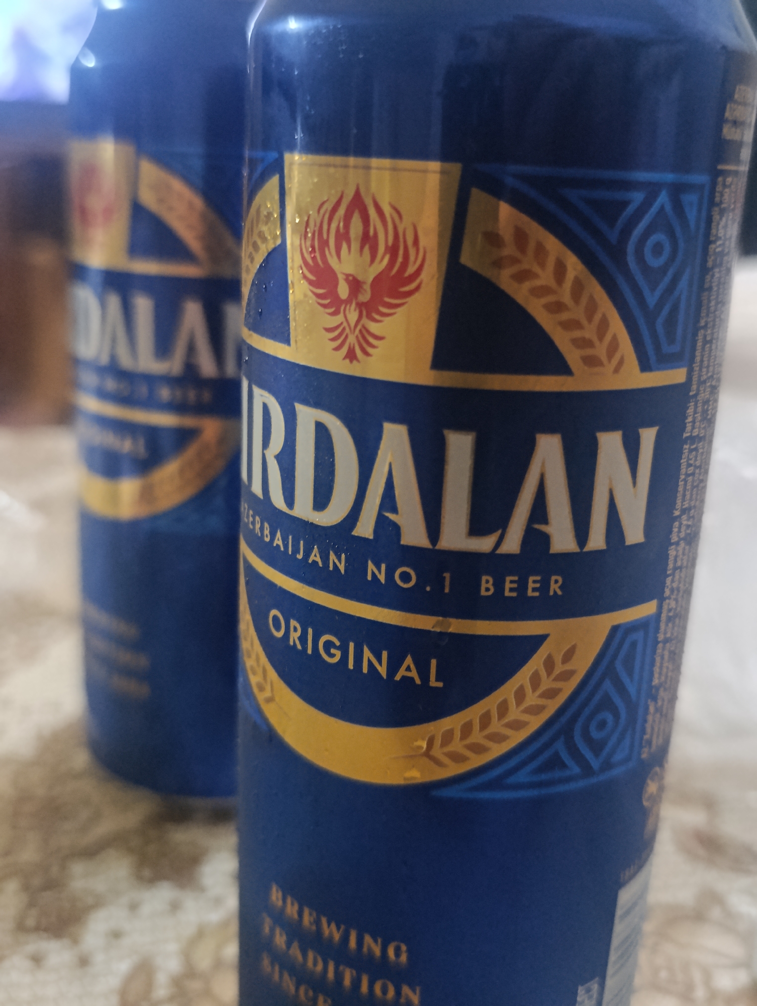The image shows two blue cans of "Xirdalan" beer. The can in the foreground is in focus and displays the brand name prominently in gold lettering. Below the brand name, "Azerbaijan No.1 Beer" and "Original" are also visible in gold. The can features a stylized red bird emblem and decorative golden wheat stalks and geometric patterns. The can in the background is out of focus but also appears to be a "Xirdalan" beer can with similar branding. The cans are on a textured surface, possibly a table, with a blurred background. There is no indication of the specific location in Baku, Azerbaijan, other than the brand being from Azerbaijan.