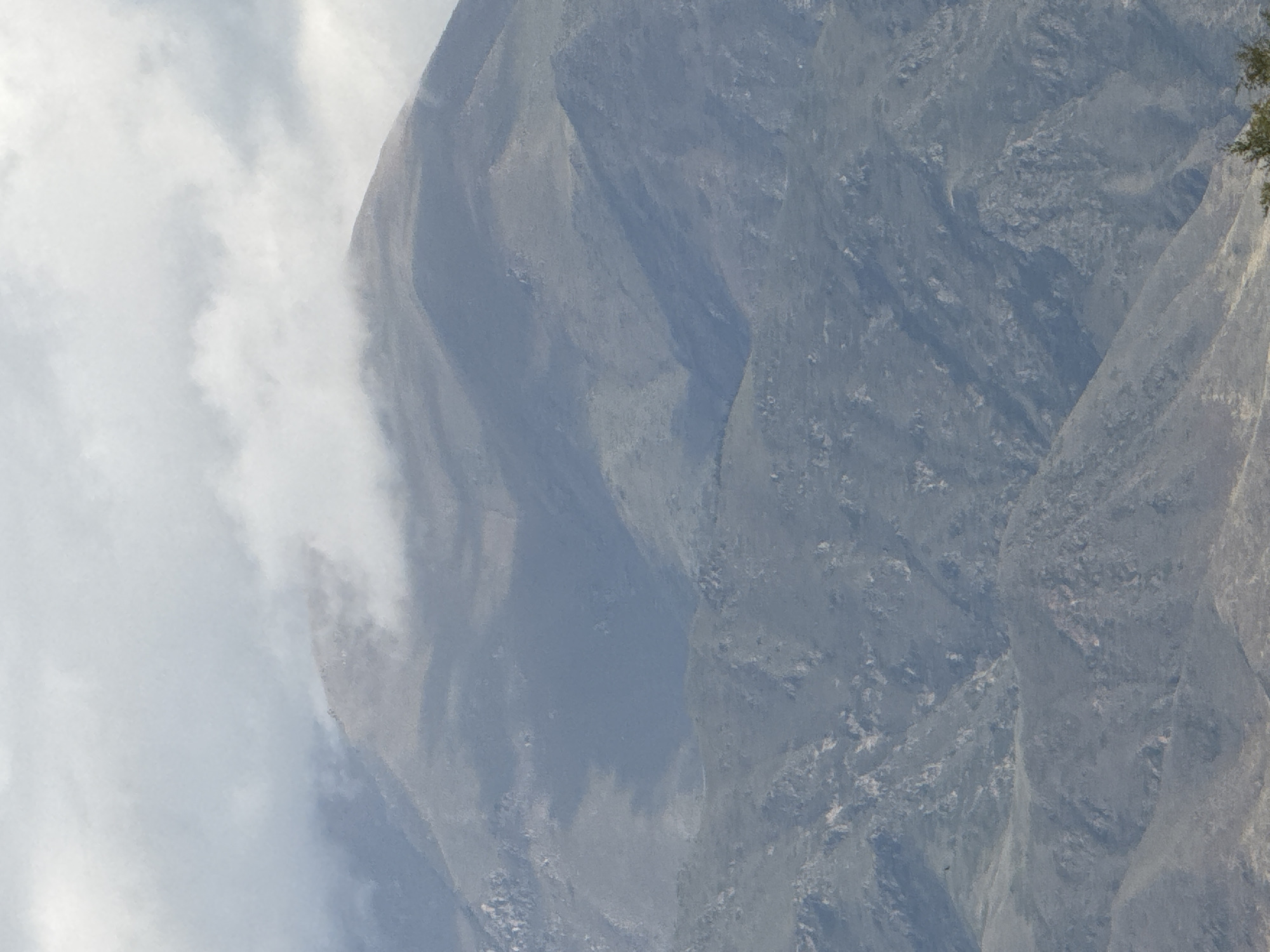 This image shows a distant, hazy view of rugged, mountainous terrain with scattered clouds obscuring the peaks. The landscape is predominantly shades of gray and blue, suggesting a cool, possibly overcast day. The texture of the mountains is rough, with visible rocky outcrops and sparse vegetation. In the lower right corner, a small portion of a green tree is visible, indicating the presence of some foliage in the foreground. No people or specific objects are clearly discernible. The scene is a natural, undeveloped environment, likely a mountainous region in or around Sary-Oy, Kyrgyzstan, given the context provided.