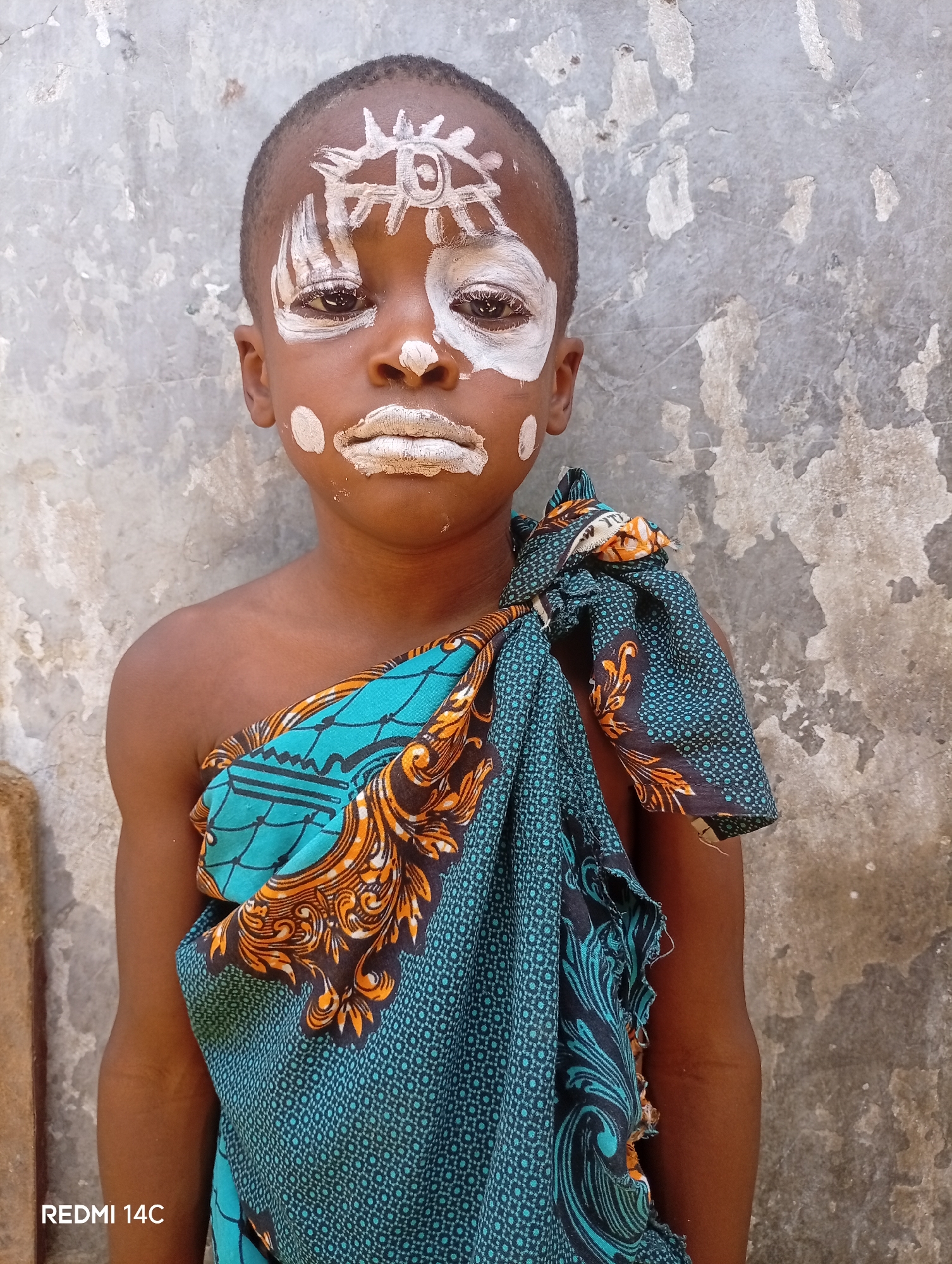 A young boy stands against a weathered wall in Maiduguri, Nigeria. He is wearing a patterned, teal and orange cloth draped over one shoulder. White paint is applied to his face in a pattern resembling a sun on his forehead, covering his eyelids, and in dots on his cheeks and lips. The wall behind him shows signs of age and wear. The photograph was taken on a Redmi 14C.