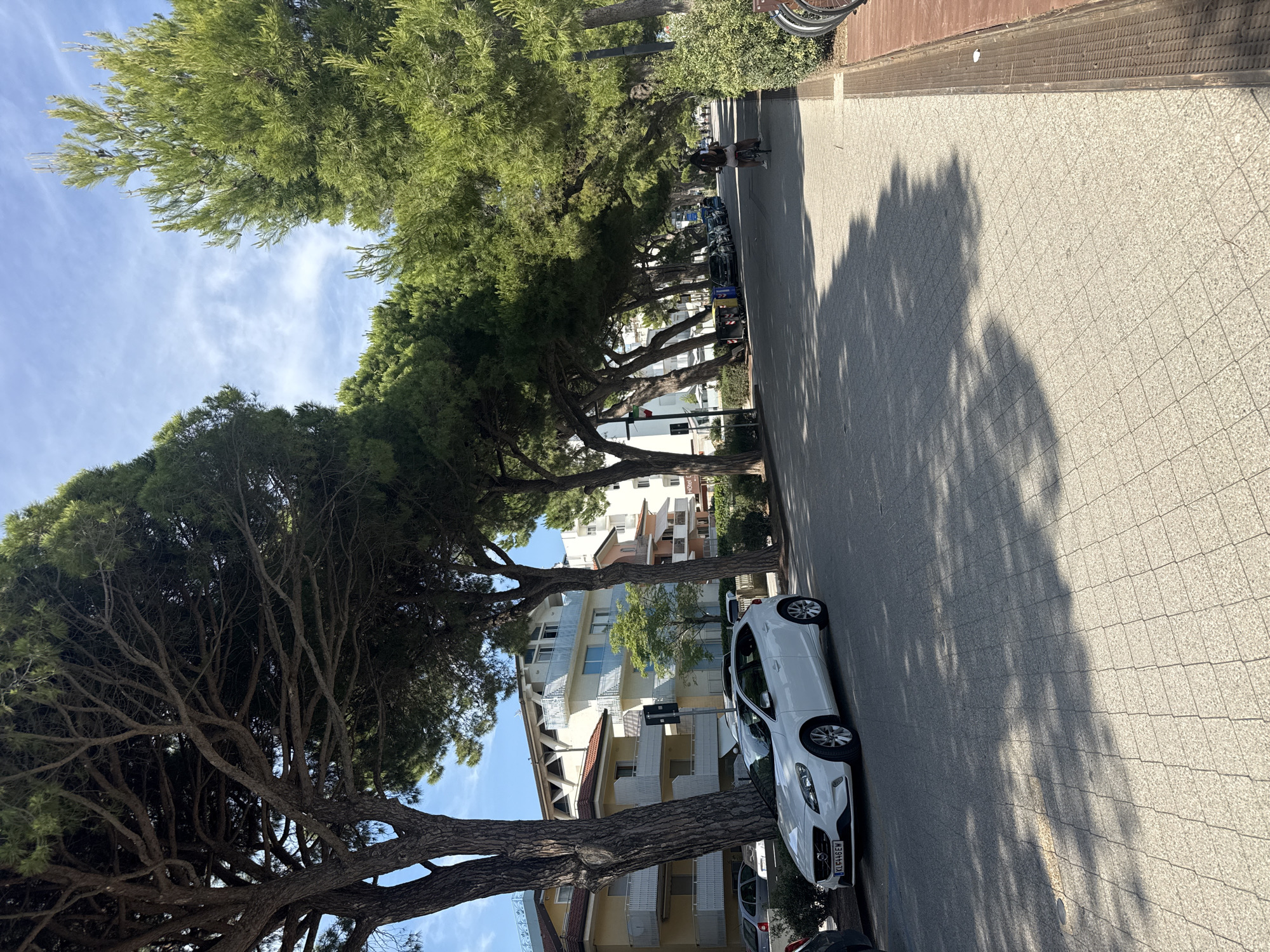 The image shows a tree-lined street in Lignano Sabbiadoro, Italy, on a sunny day. A white Volvo V40 is parked on the left side of the street, with its license plate reading "KLE446EW". A woman is cycling on the right side of the street, and there are several buildings and trees visible in the background. The street is paved with light-colored tiles, and shadows from the trees are cast across the pavement. In the distance, more parked cars and a few people can be seen. The overall scene depicts a tranquil urban environment with abundant greenery.