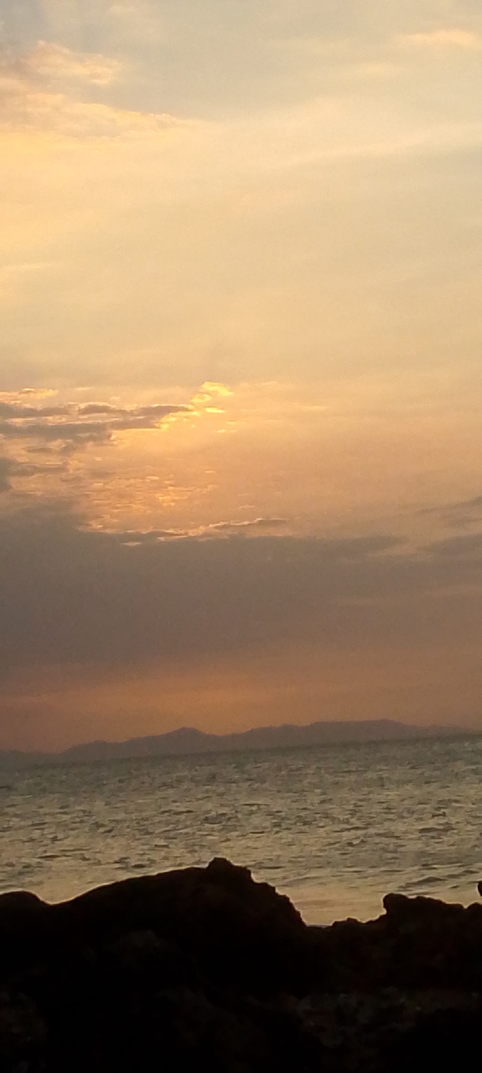 This image depicts a sunset over a body of water, likely the Caribbean Sea, viewed from a rocky shoreline in Juan Griego, Venezuela. The upper two-thirds of the frame are occupied by a sky exhibiting a gradient of colors from pale yellow-orange at the top to deeper orange and pink hues towards the horizon. Scattered clouds, partially obscuring the sun in the upper left-center, allow golden light rays to penetrate. A dark, undulating silhouette of distant mountains or hills stretches across the central horizon line. Below this, the ocean surface appears textured with small ripples, reflecting the ambient light. The immediate foreground, comprising the bottom one-fifth of the image, is dominated by dark, irregularly shaped rock formations, presented as a complete silhouette against the brighter background. No human figures, animals, structures, or discernible objects of human origin are present within the scene.