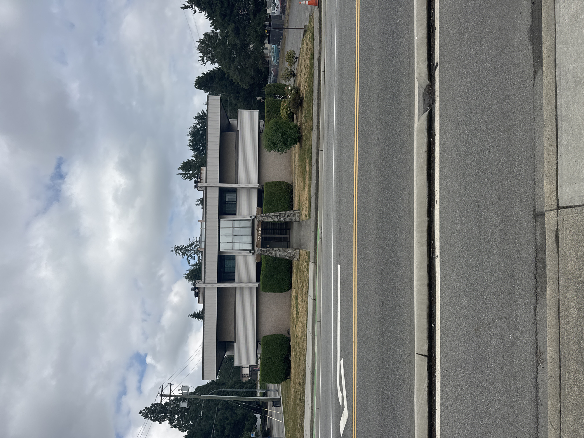 An exterior, eye-level, medium-shot photograph captures a two-story, light-beige, modern apartment building situated in Surrey, Canada. The building has a flat roof and is characterized by horizontal siding and protruding balconies. The entrance is centrally located and framed by two stone pillars, with the address "10188" visible above the doorway. The building is fronted by a lawn and two symmetrical, rounded, dark-green bushes on either side of the entrance, with a pathway leading to the building. Across the street, a gray asphalt road displays yellow lane markings and a white turn arrow. In the background, mature evergreen trees line the property, and a few parked cars are visible. The sky is overcast with clouds.