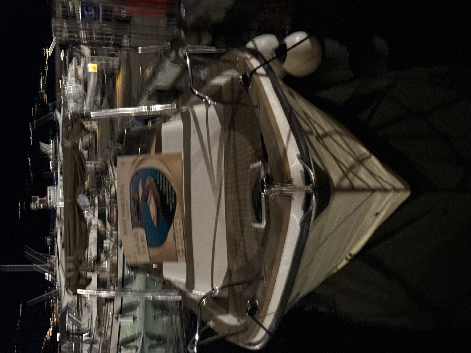 A motorboat is docked at night in Budva, Montenegro, with other boats and lights in the background. The boat has a white hull with wooden accents and chrome railings. A sign with an image of a smaller boat is displayed on the front of the boat. Two white fenders are attached to the side of the boat. The water is dark, and reflections of the boat and other lights are visible.